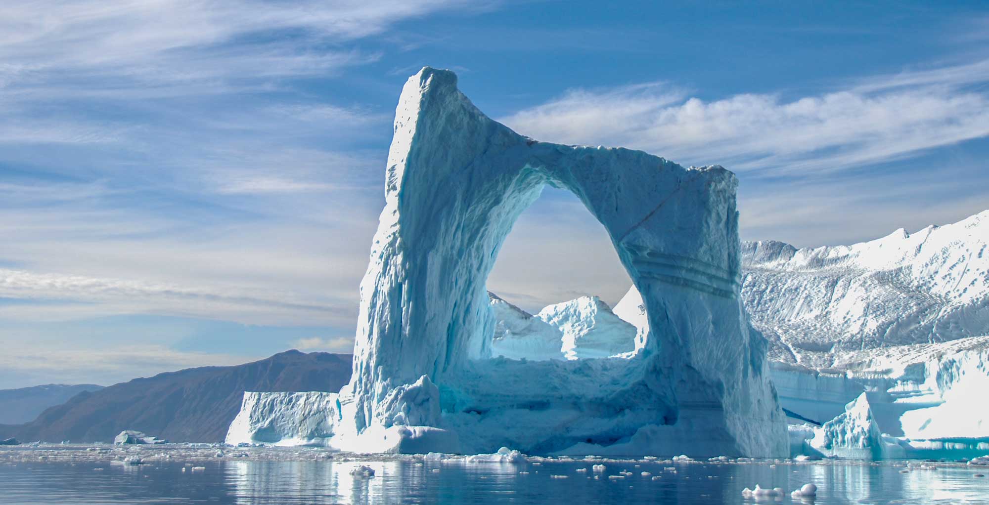 Massive blue iceberg with natural arch opening, floating in calm waters near snow-capped mountains