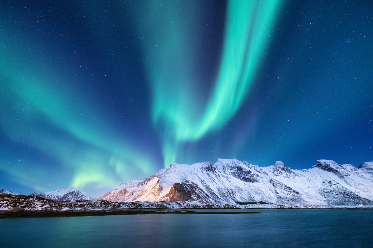 Spectacular aurora borealis dancing in vibrant green and blue hues above snow-covered mountains and a calm fjord waters.