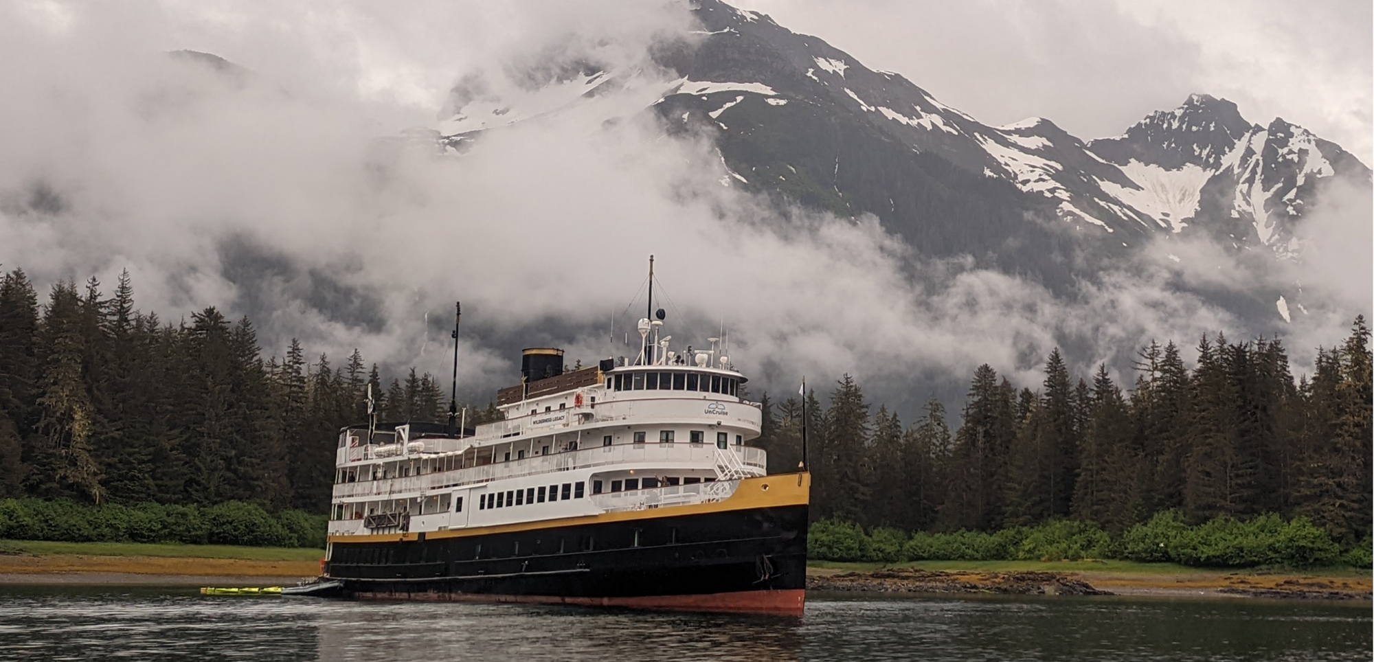 Alaskan Ferry in Misty Mountain Landscape A white and yellow passenger ferry navigates through calm waters near a forested shoreline