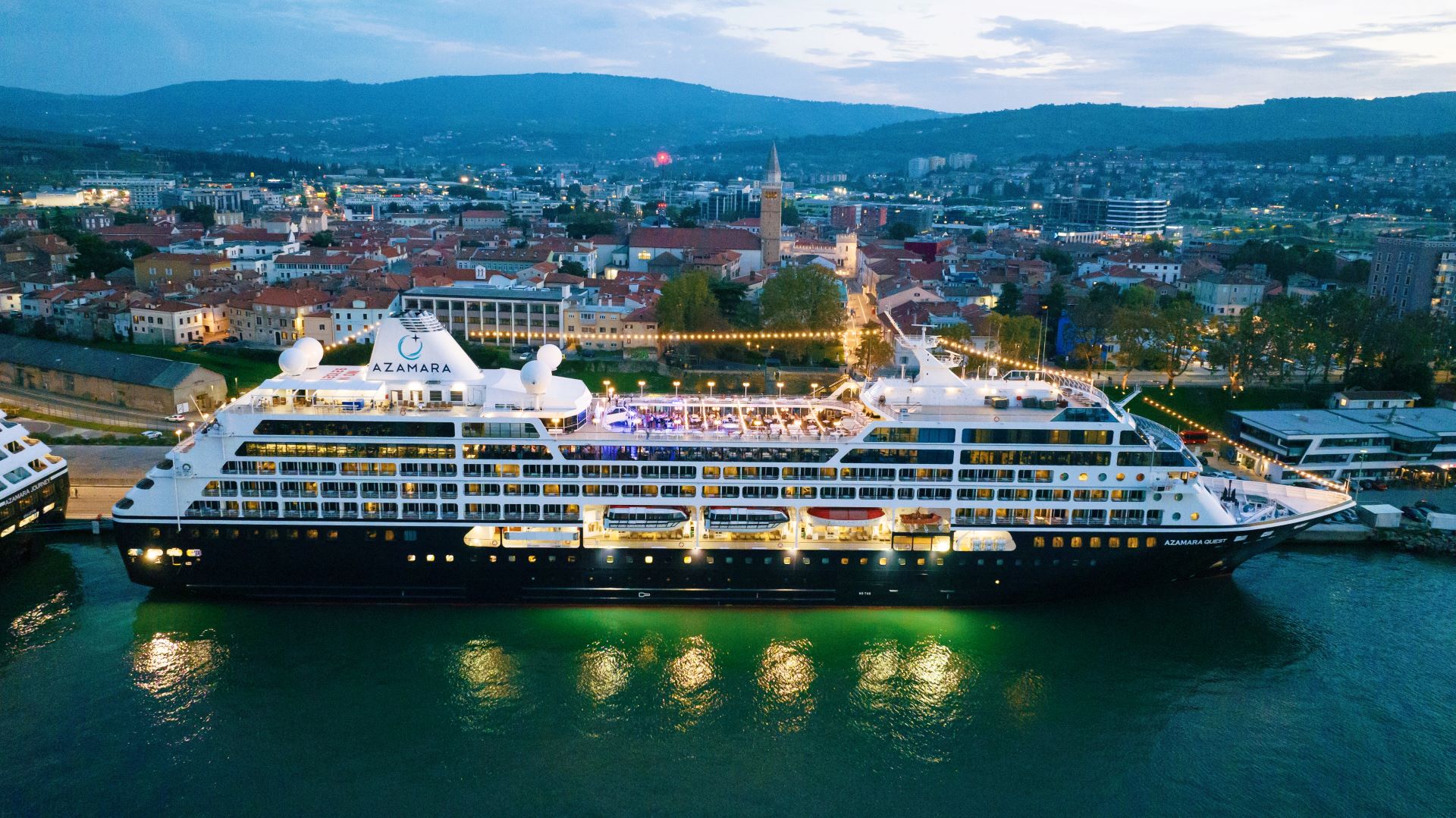 An Azamara cruise ship illuminated at night, docked in a picturesque European city with mountains and urban landscape in the background.