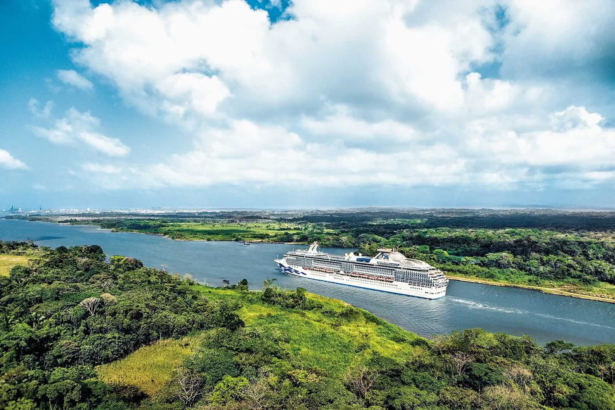 A large modern cruise ship navigates through Gatun Lake surrounded by lush tropical rainforest and verdant vegetation. The image captures an aerial perspective of the vessel in the waterway with dense green foliage on both shorelines and a bright blue sky with white clouds overhead.