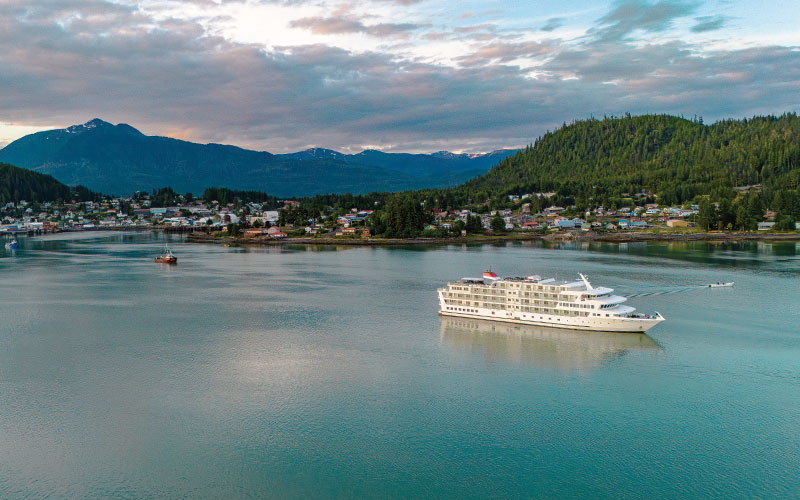 Cruise Ship in Alaskan Coastal Town A white cruise ship anchored in a tranquil bay near a forested coastal town with mountains in the background