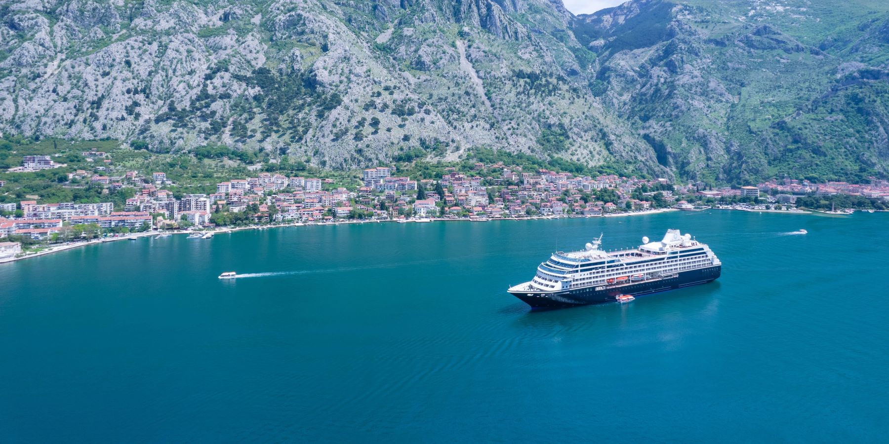 A large cruise ship anchored in a turquoise bay surrounded by steep, rocky mountains with