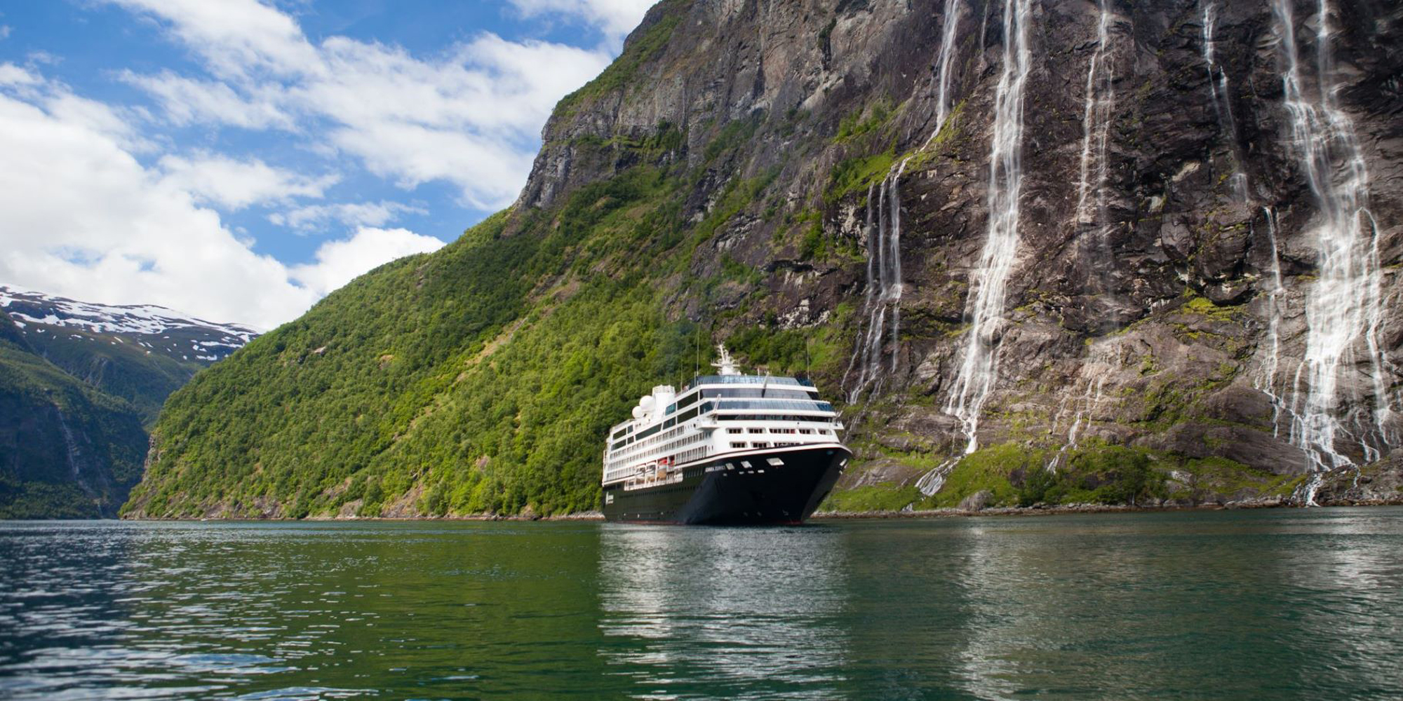 A white cruise ship sailing through a dramatic fjord landscape with steep, green-covered mountains and multiple waterfalls cascading down rocky cliffs.