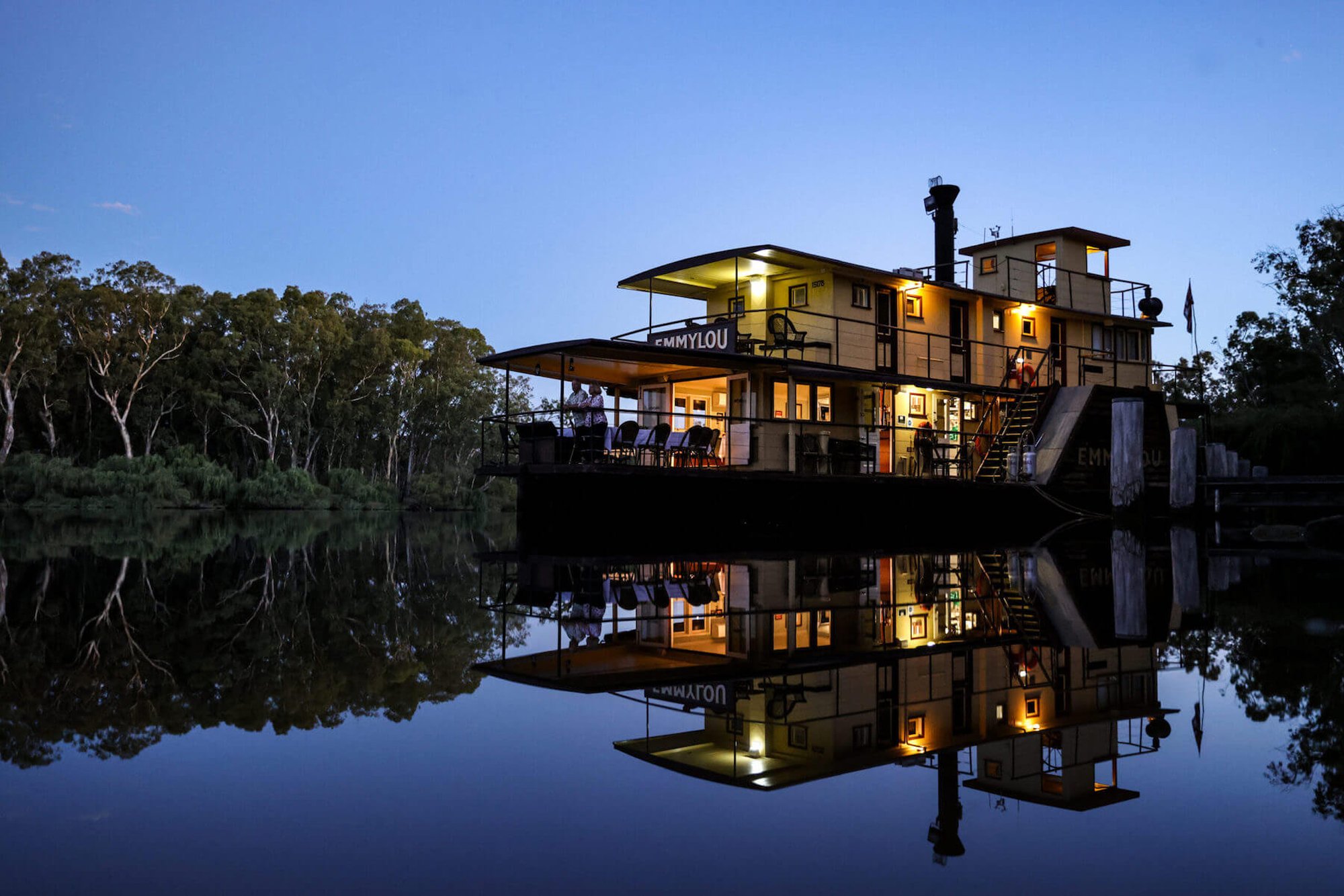 A beautifully illuminated multi-level houseboat named Emmylou glows warmly against the twilight sky, its reflection perfectly mirrored in the still, dark waters below. Surrounded by eucalyptus trees along a serene riverbank, the vessel showcases modern architecture with multiple decks, large windows, and outdoor living spaces.