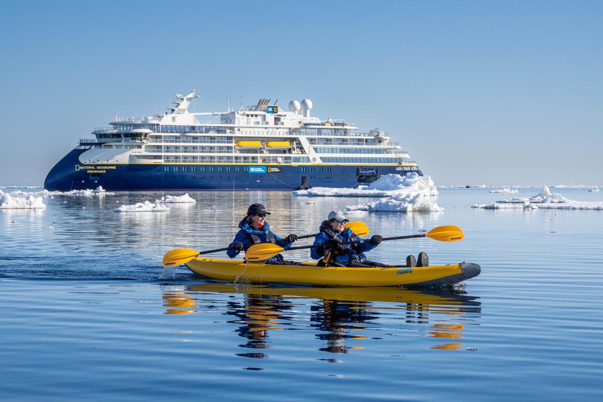 Two kayakers paddle in a bright yellow kayak in pristine Arctic waters surrounded by icebergs