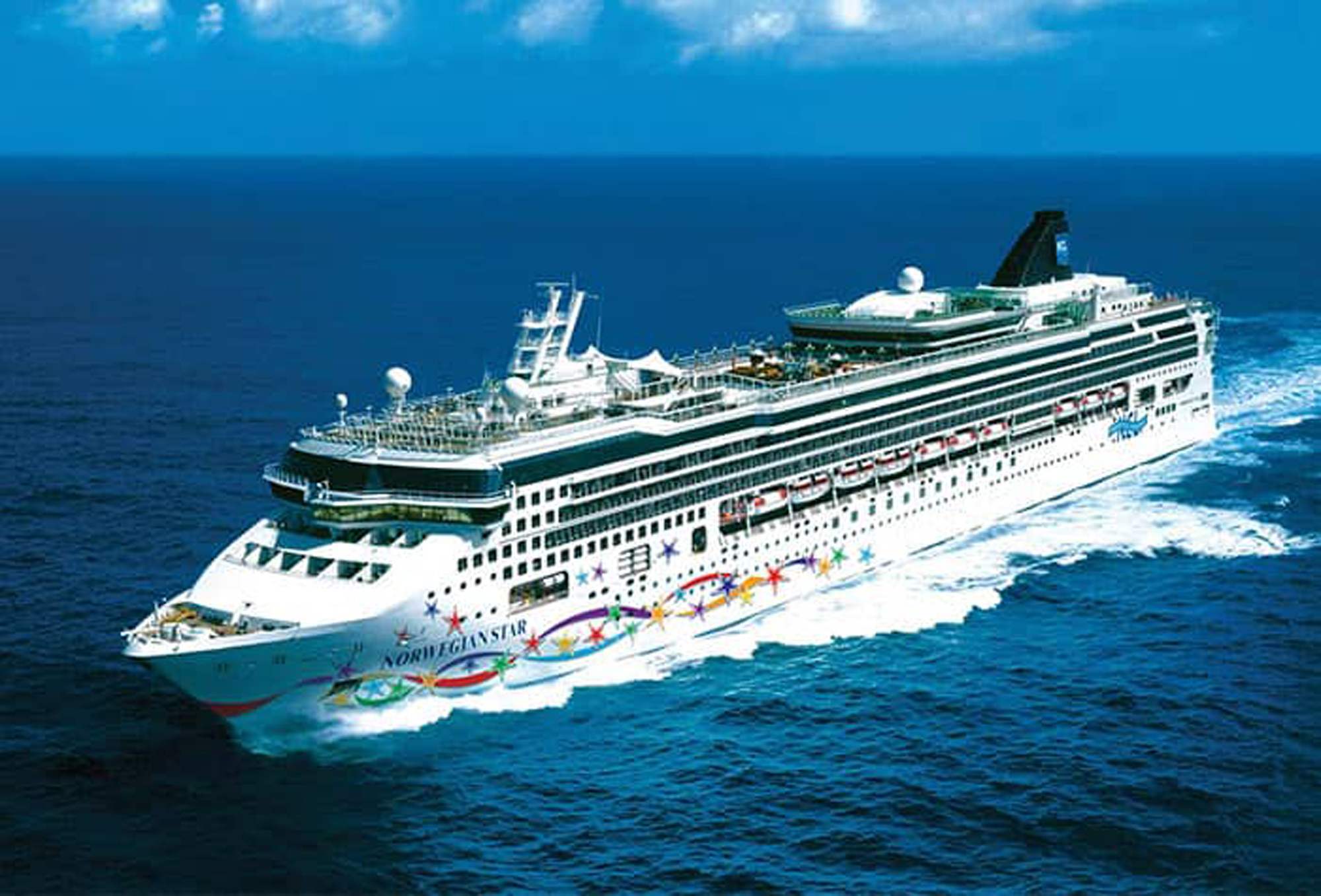A large white Norwegian Star cruise ship sailing through deep blue ocean waters, decorated