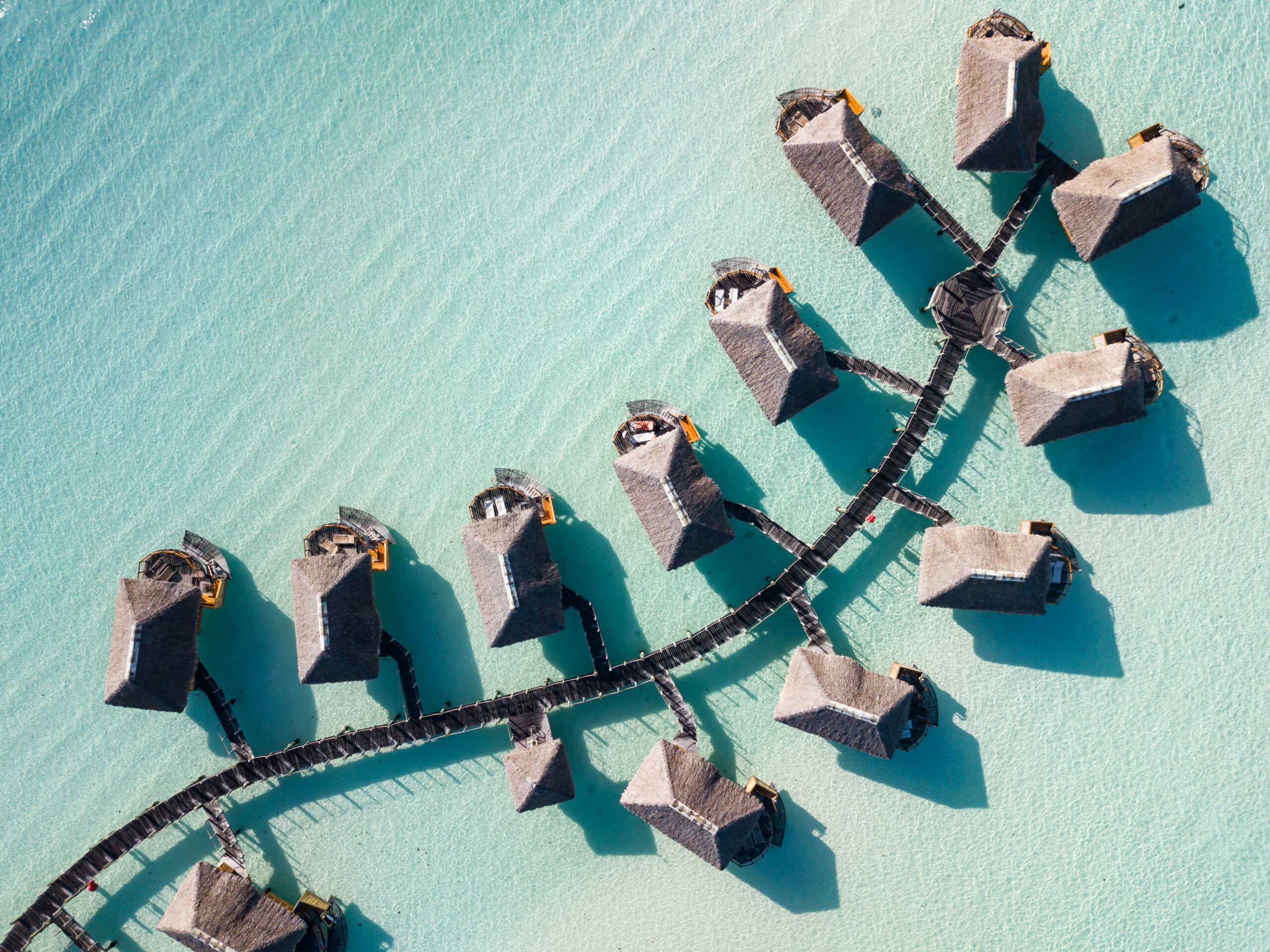 Overwater Bungalows Aerial View An aerial view of a luxury resort with thatched-roof overwater bungalows arranged in a curved pattern along wooden walkways in crystal-clear turquoise lagoon waters. The image showcases the pristine tropical destination with perfect symmetry and shallow waters characteristic of South Pacific island resorts.