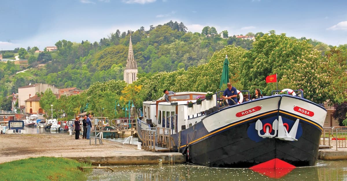 The river cruise ship Rosa is moored at a picturesque European riverport with passengers on deck, surrounded by lush green hillsides, a church spire visible in the background, and traditional riverside architecture. The scene captures the essence of leisurely river cruising through a charming continental landscape.