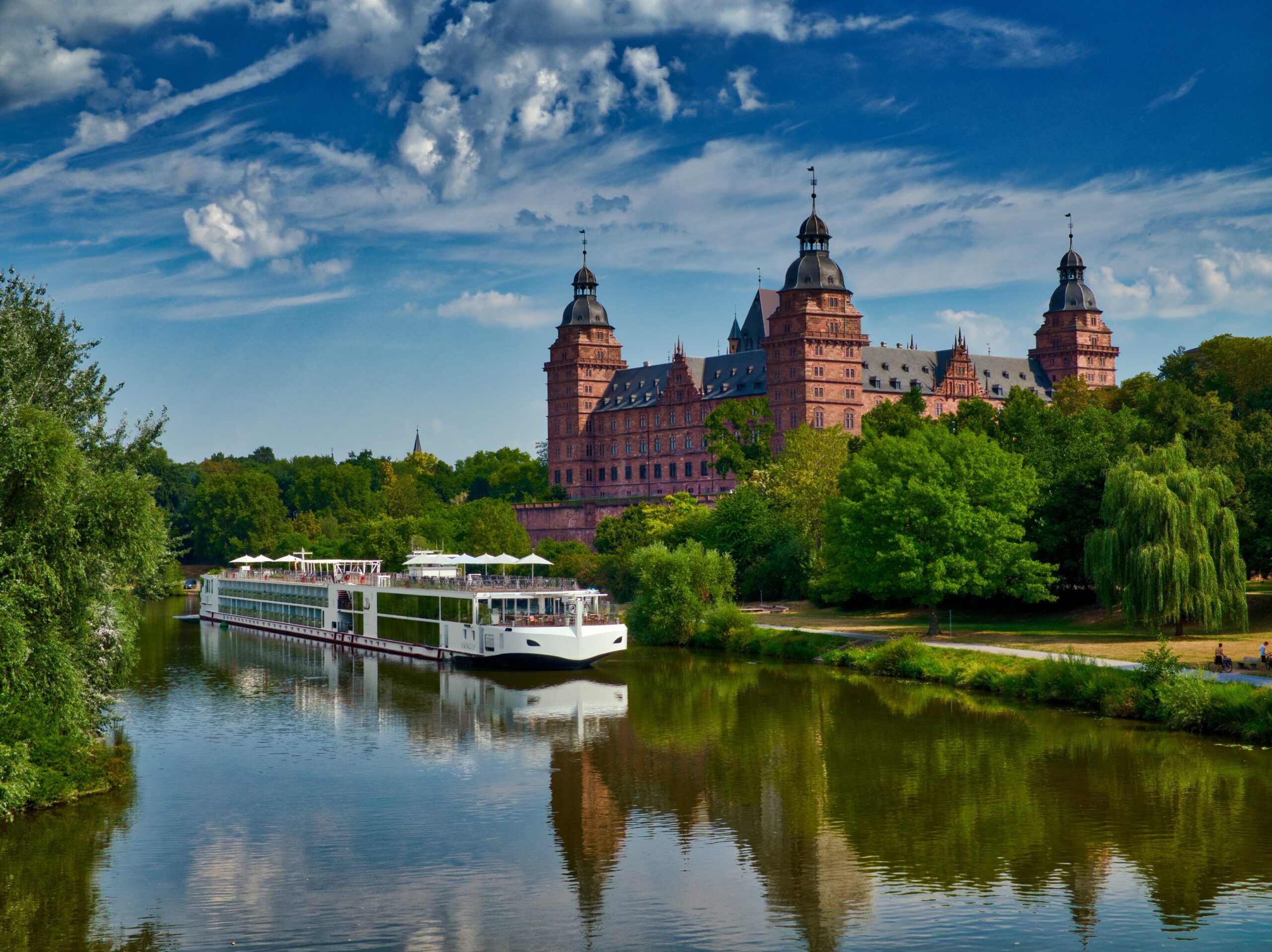 River Cruise Past Historic German Castle A modern river cruise ship navigates a tranquil river in front of an impressive Renaissance-style castle with multiple towers, surrounded by lush green trees.