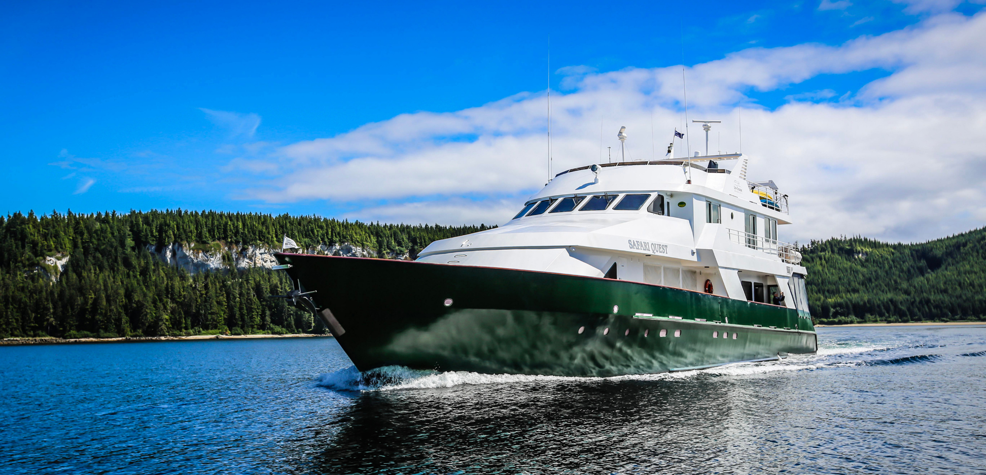 Safari Quest Expedition Vessel in Forested Waterway A white and green expedition cruise ship named Safari Quest navigating through a scenic forested