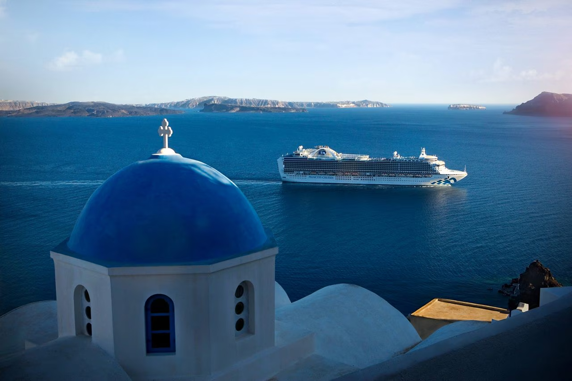 A classic white and blue church dome overlooks a large cruise ship anchored in the deep blue waters
