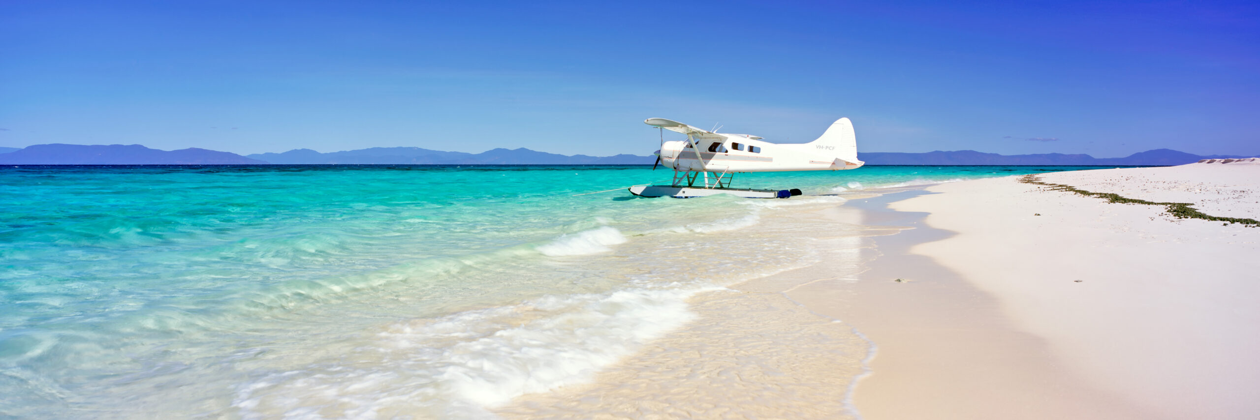 A white twin-engine seaplane is moored in the crystal-clear turquoise waters of a pristine tropical beach, with powdery white sand and calm shallow waters stretching to the shoreline. Mountains are visible across the horizon under a clear blue sky, suggesting a remote island destination perfect for scenic flights and water-based tourism.