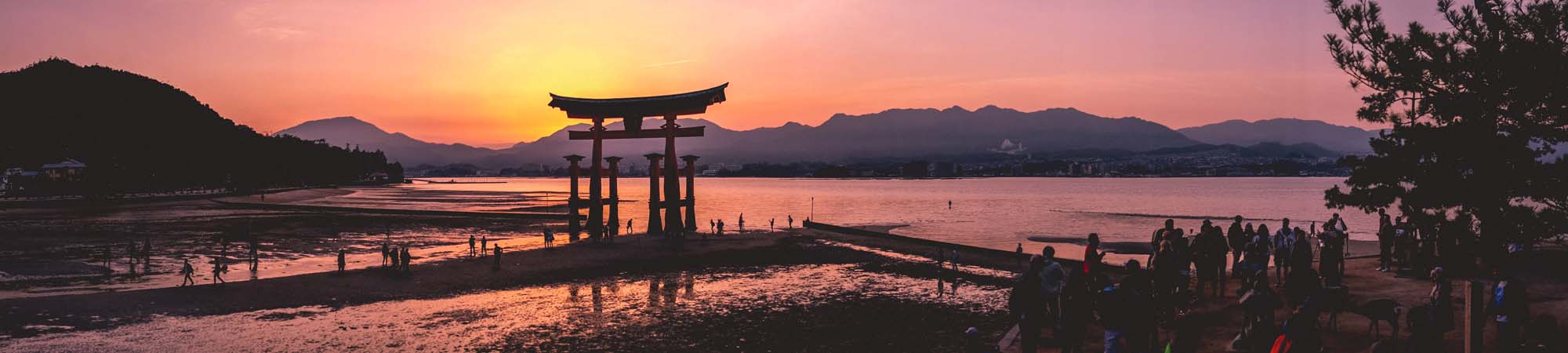 A stunning sunset view of the iconic Itsukushima Shrine floating torii gate at low tide, with silhouetted visitors and mountains in the background.