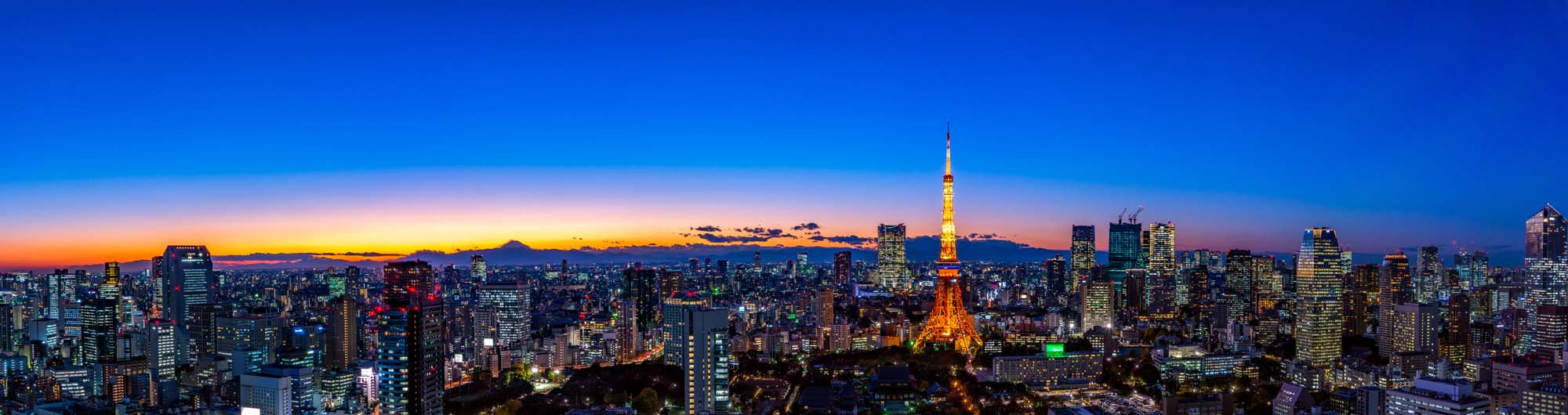 A breathtaking panoramic view of Tokyo's urban landscape at sunset, featuring the iconic Tokyo Tower illuminated against a gradient sky with Mount Fuji in the distant background.