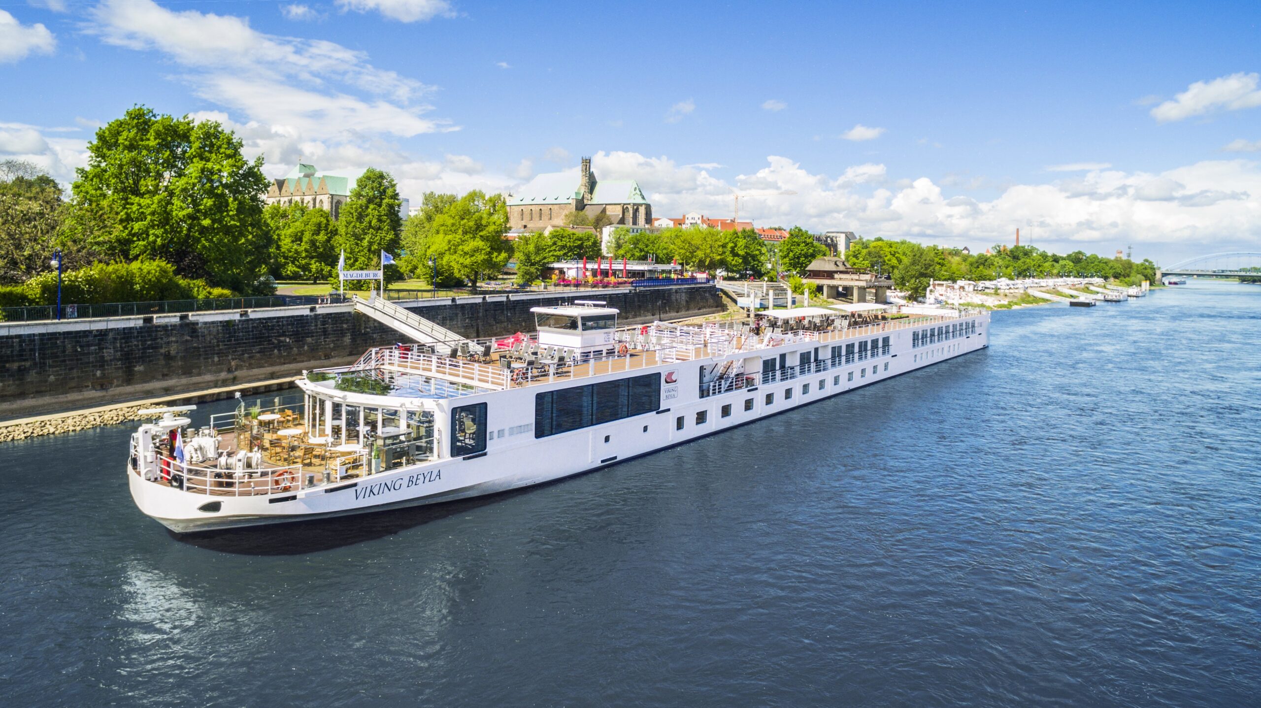 Viking River Cruise on European Waterway A white Viking river cruise ship docked near a historic riverside city with green trees and architectural landmarks.