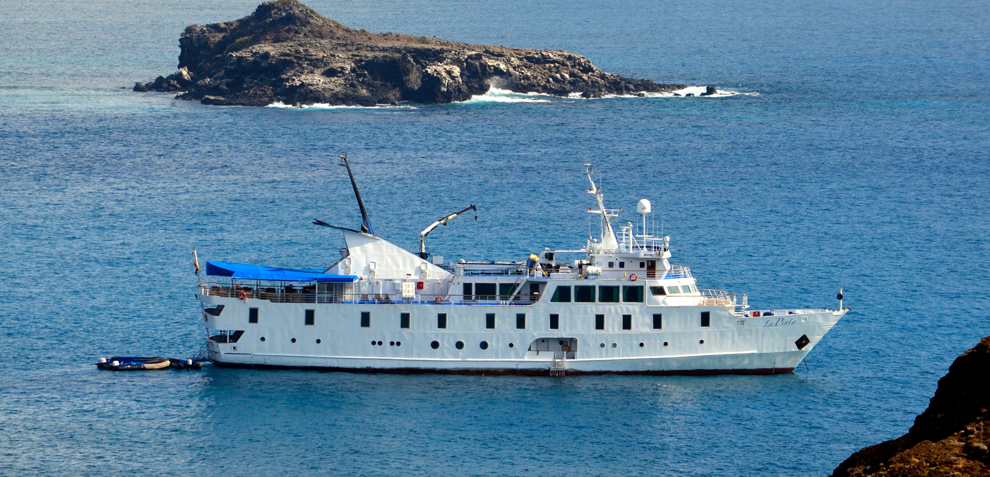 White Cruise Ship Near Rocky Island White expedition cruise vessel anchored near a rocky volcanic island with blue ocean waters