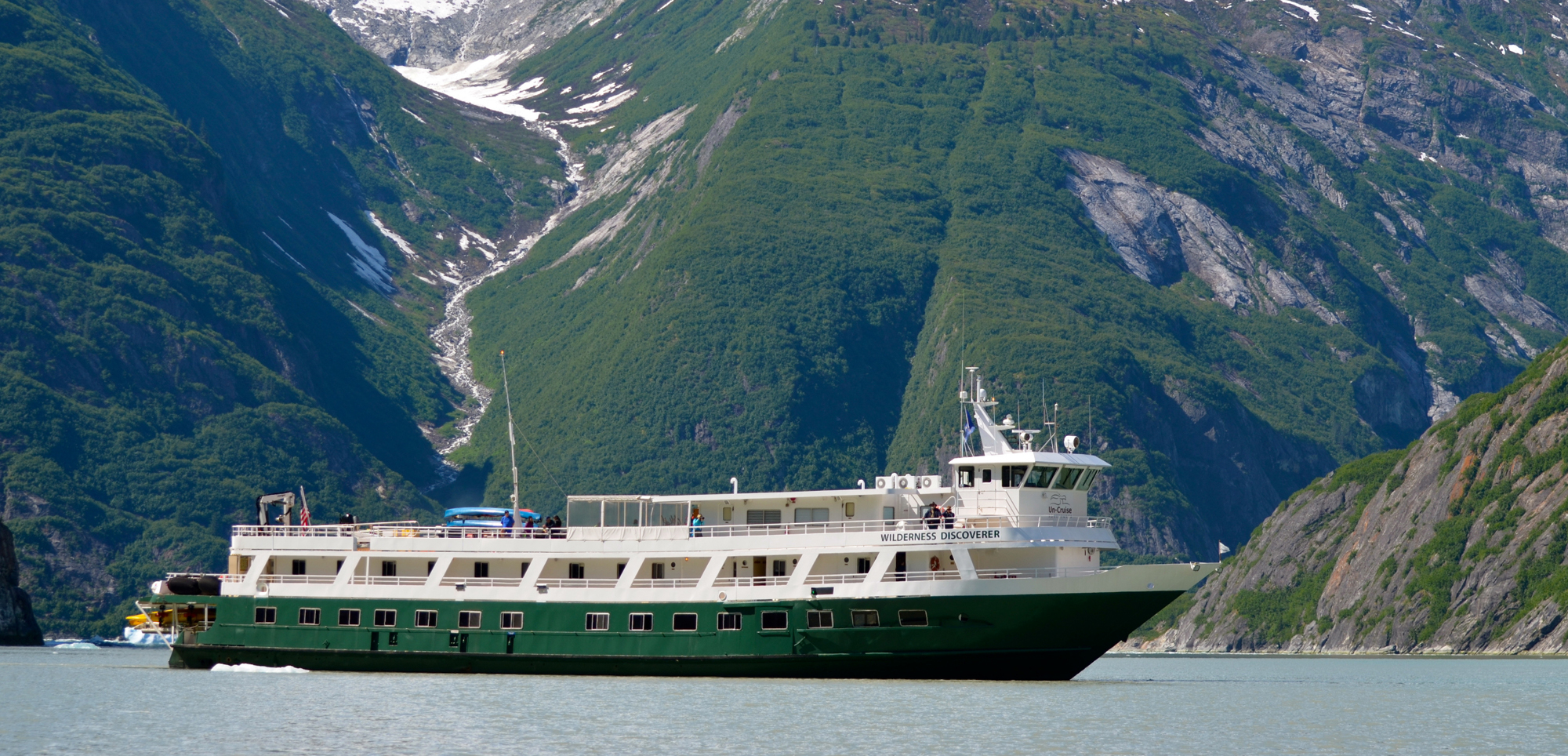 A green and white expedition cruise ship navigating through a stunning fjord with steep, forested