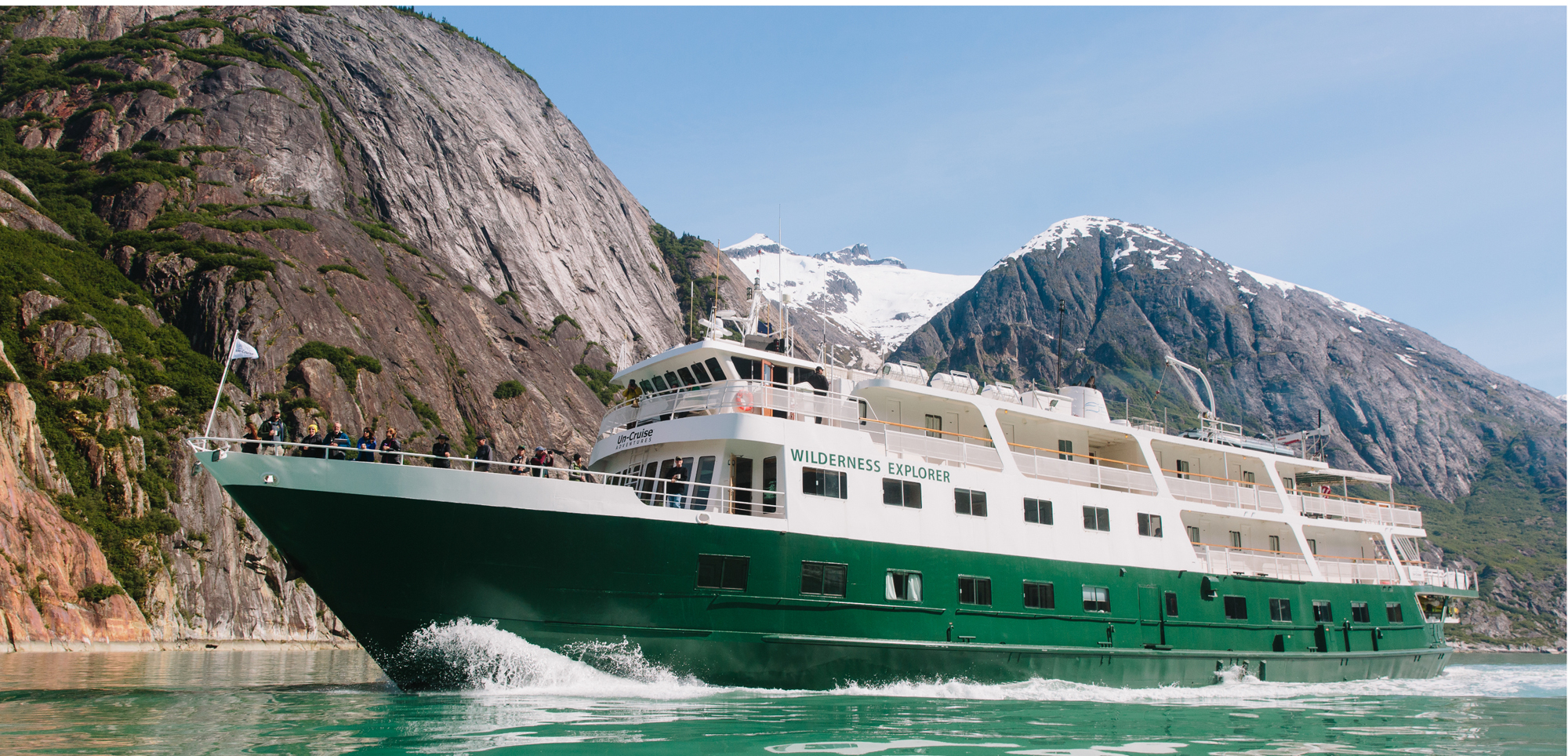 Wilderness Explorer Cruise in Alaska A green and white expedition cruise ship navigating through a scenic Alaskan fjord with dramatic