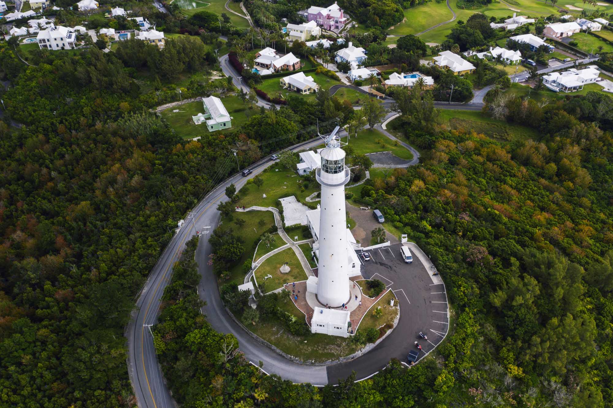 Aerial View of Bermuda Lighthouse and Coastal Community An aerial photograph of a distinctive white lighthouse with its accompanying keeper's residence situated on a promontory in Bermuda, surrounded by lush green vegetation, winding coastal roads, and residential properties.