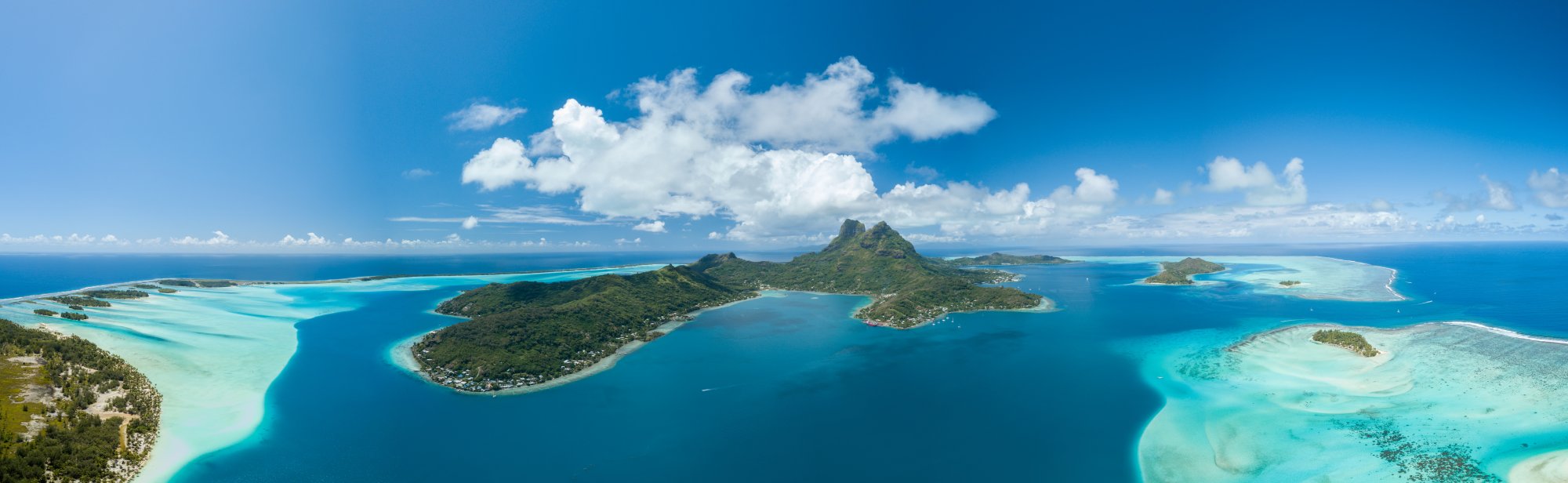 A stunning aerial panorama of Bora Bora, French Polynesia, showcasing the dramatic twin volcanic peaks rising from lush green vegetation, surrounded by crystal-clear turquoise lagoon waters with vibrant coral reef patterns visible in the shallow areas, dotted with small islands and settlements.