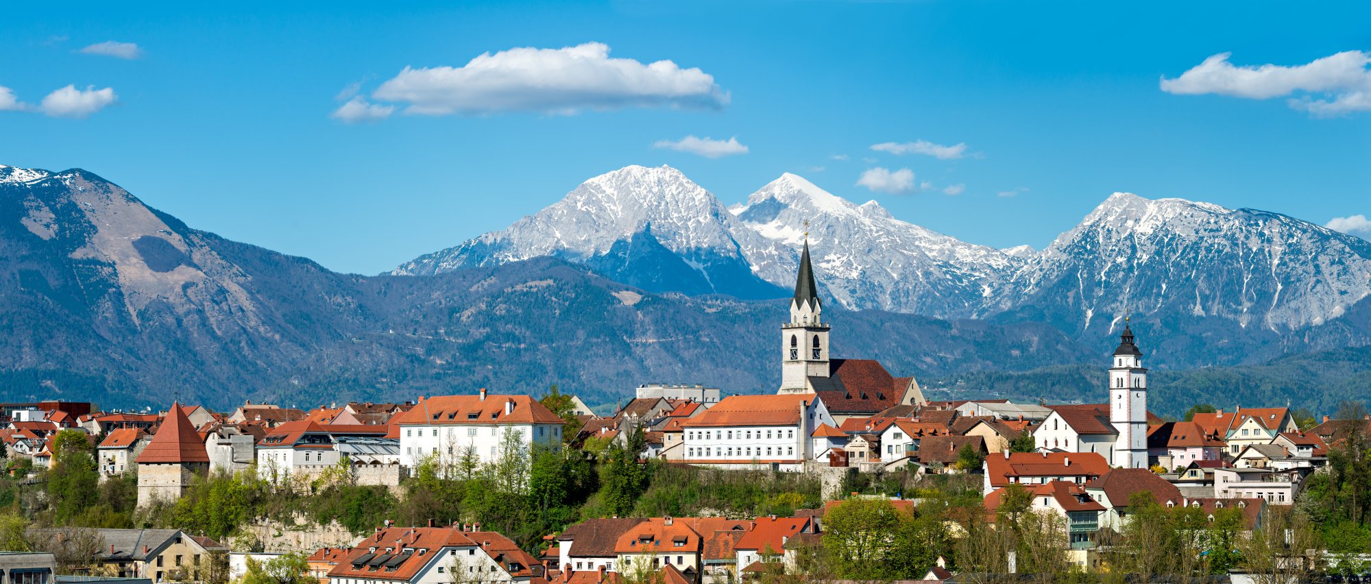 A picturesque Alpine village nestled in a valley with distinctive orange-tiled roofs and white church spires, framed by dramatic snow-covered mountain peaks in the background under a clear blue sky. The landscape exemplifies the natural beauty and architectural charm of the Bavarian Alps region.