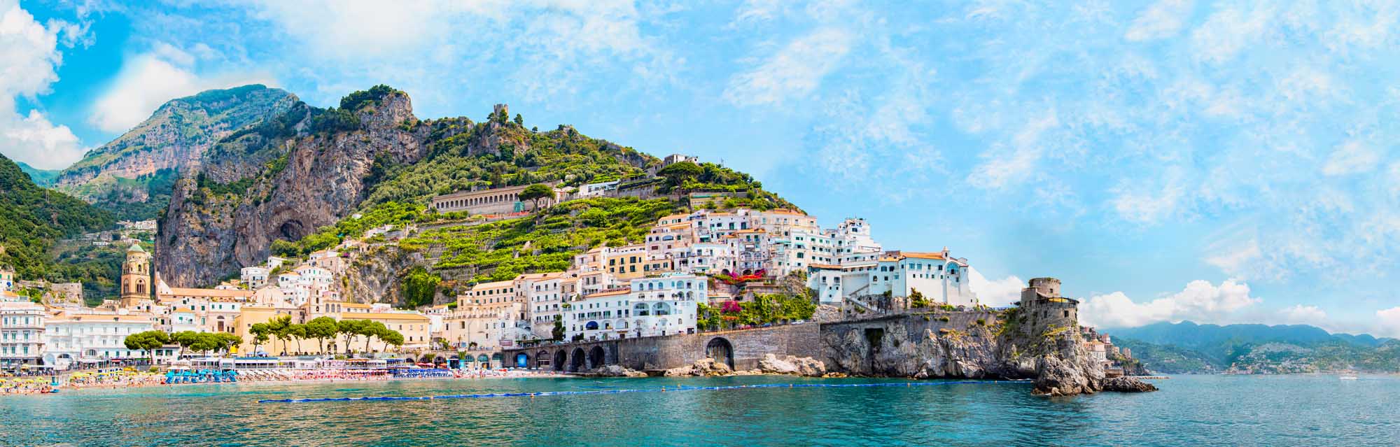Amalfi Village Coastal Panorama Italy A stunning aerial panoramic view of the picturesque Amalfi village perched on dramatic rocky cliffs along the Amalfi Coast in Italy. The scene showcases colorful pastel-hued buildings cascading down the mountainside, a small sandy beach below, turquoise Mediterranean waters, and lush green vegetation covering the surrounding peaks under a bright blue sky.