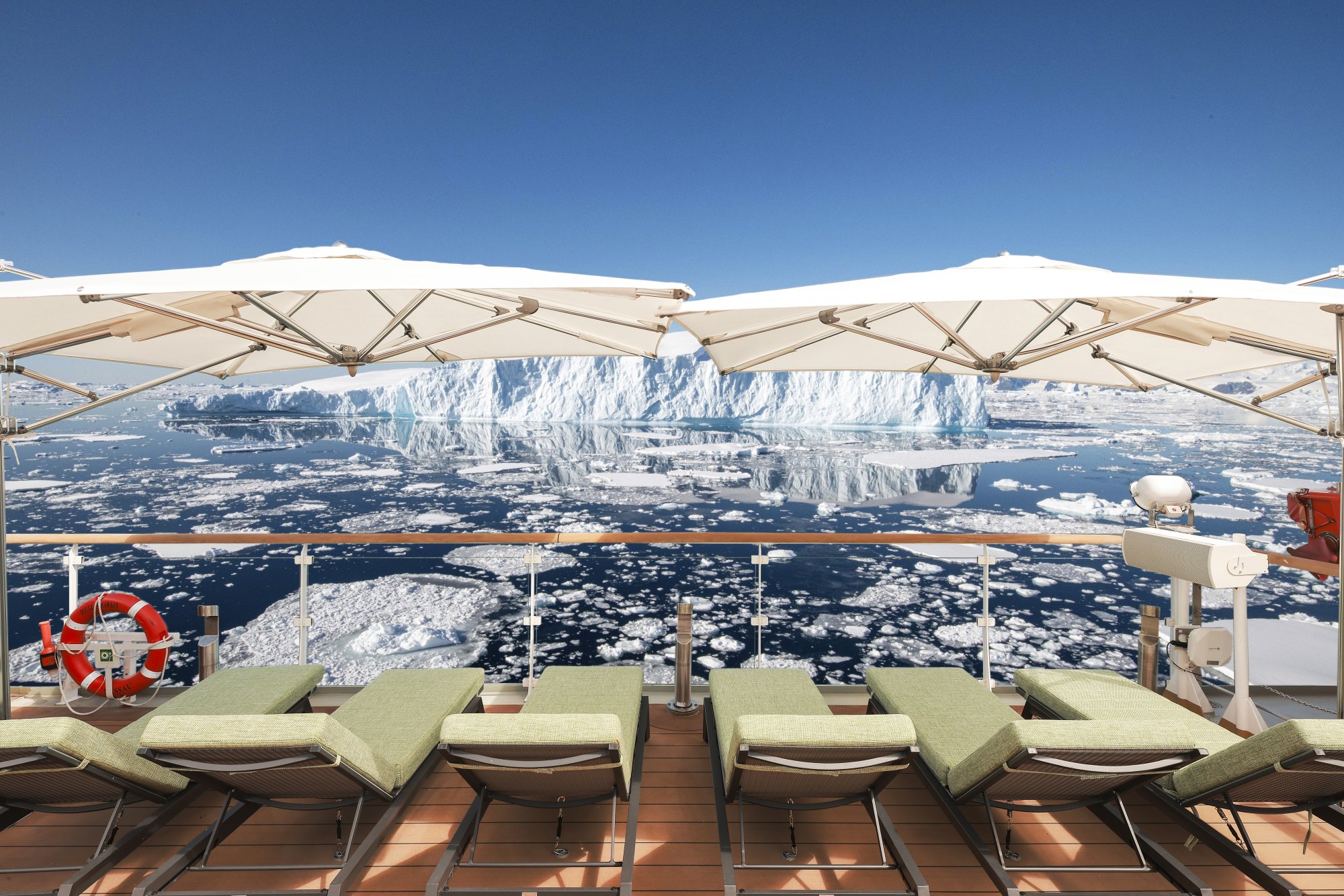 Arctic Expedition Ship Sun Deck with Glaciers A luxurious sun deck aboard an expedition cruise ship featuring comfortable loungers with green cushions under cream umbrellas, overlooking a pristine Arctic seascape dotted with icebergs and a massive glacier calving wall in the distance under clear blue skies.