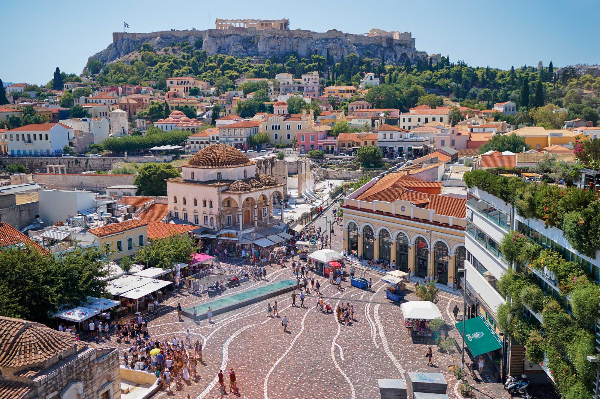 Athens Skyline with Acropolis and Monastiraki Square A vibrant daytime aerial view of Athens' historic Monastiraki Square featuring the distinctive domed mosque, colorful neoclassical buildings, bustling marketplace, and the iconic Acropolis perched dramatically on the hilltop in the background under clear blue skies.
