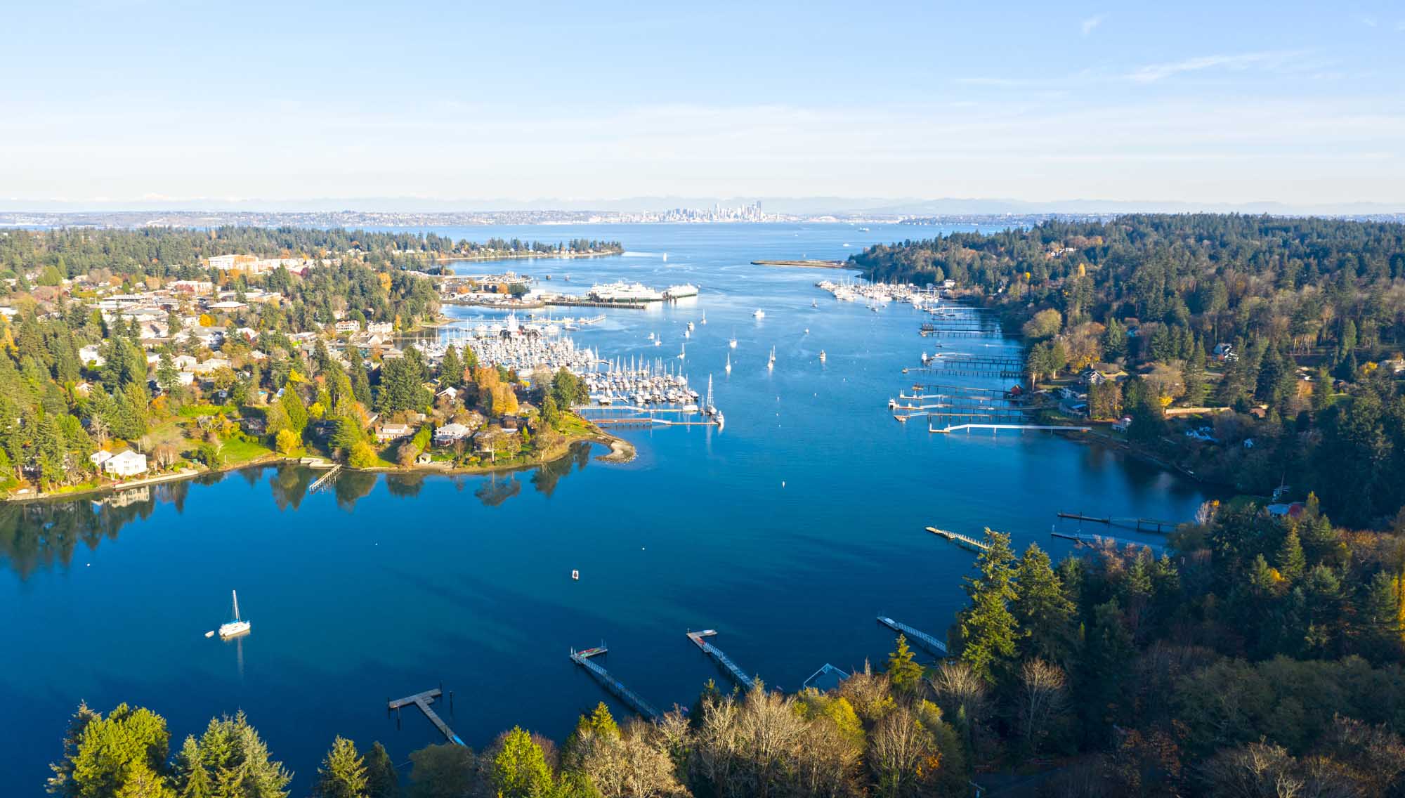 An aerial panoramic view of Bainbridge Island's scenic harbor in Washington State, featuring sailboats and moored vessels in the crystal-clear blue water, surrounded by dense evergreen forests and waterfront residential properties, with Seattle's skyline visible in the distant background across Puget Sound.