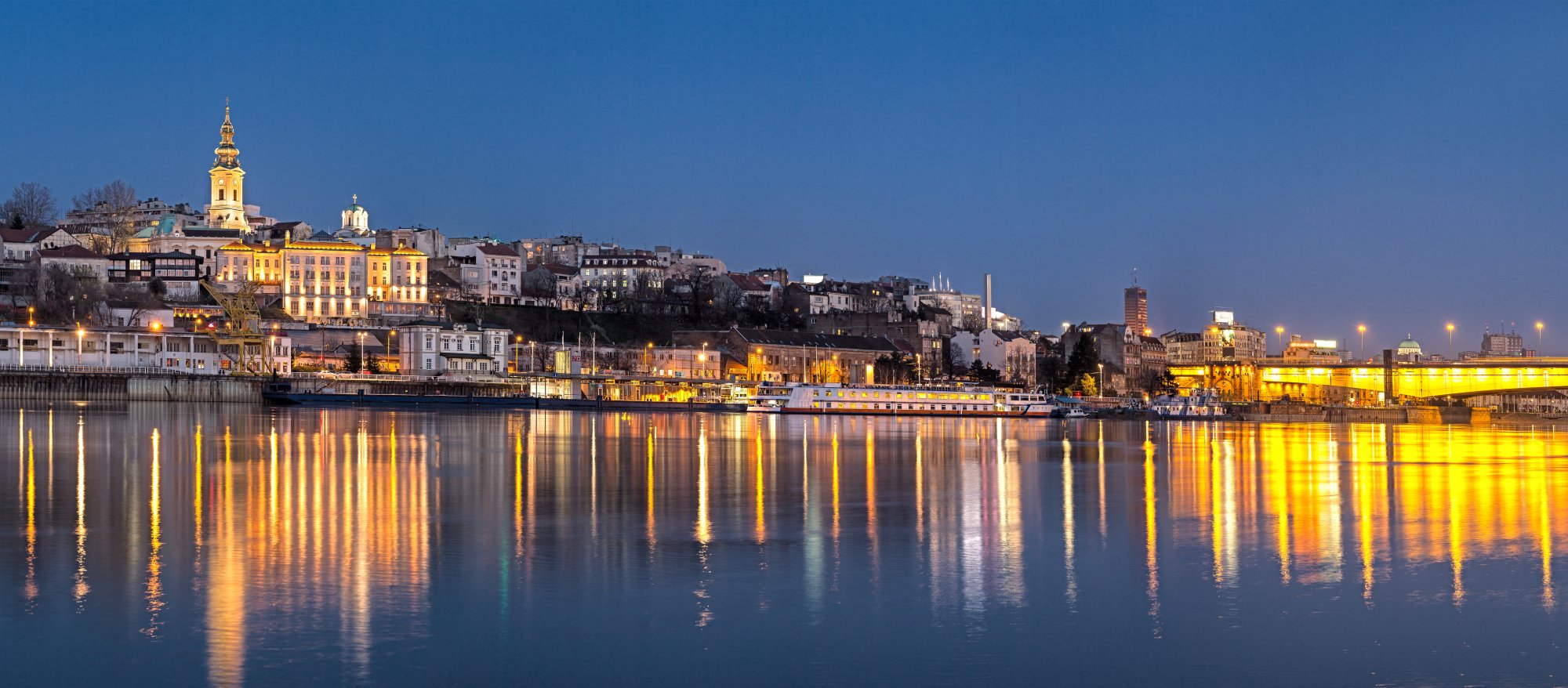 A stunning twilight view of Belgrade's Danube riverfront, featuring illuminated historic architecture reflected in calm waters. The golden-lit buildings, distinctive church spires, and riverside promenade showcase the city's architectural charm and vibrant nightlife along the Danube River.