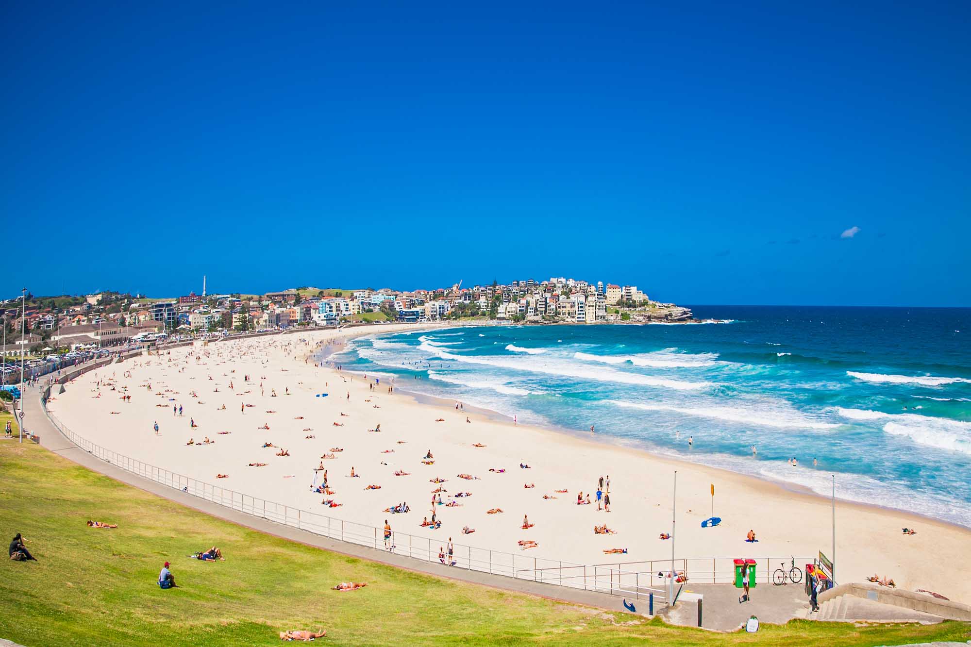 A vibrant aerial view of Bondi Beach in Sydney, Australia, featuring golden sand packed with beachgoers, turquoise ocean waves, and the iconic beachside suburb with colorful residential buildings cascading up the hillside under a brilliant blue sky.