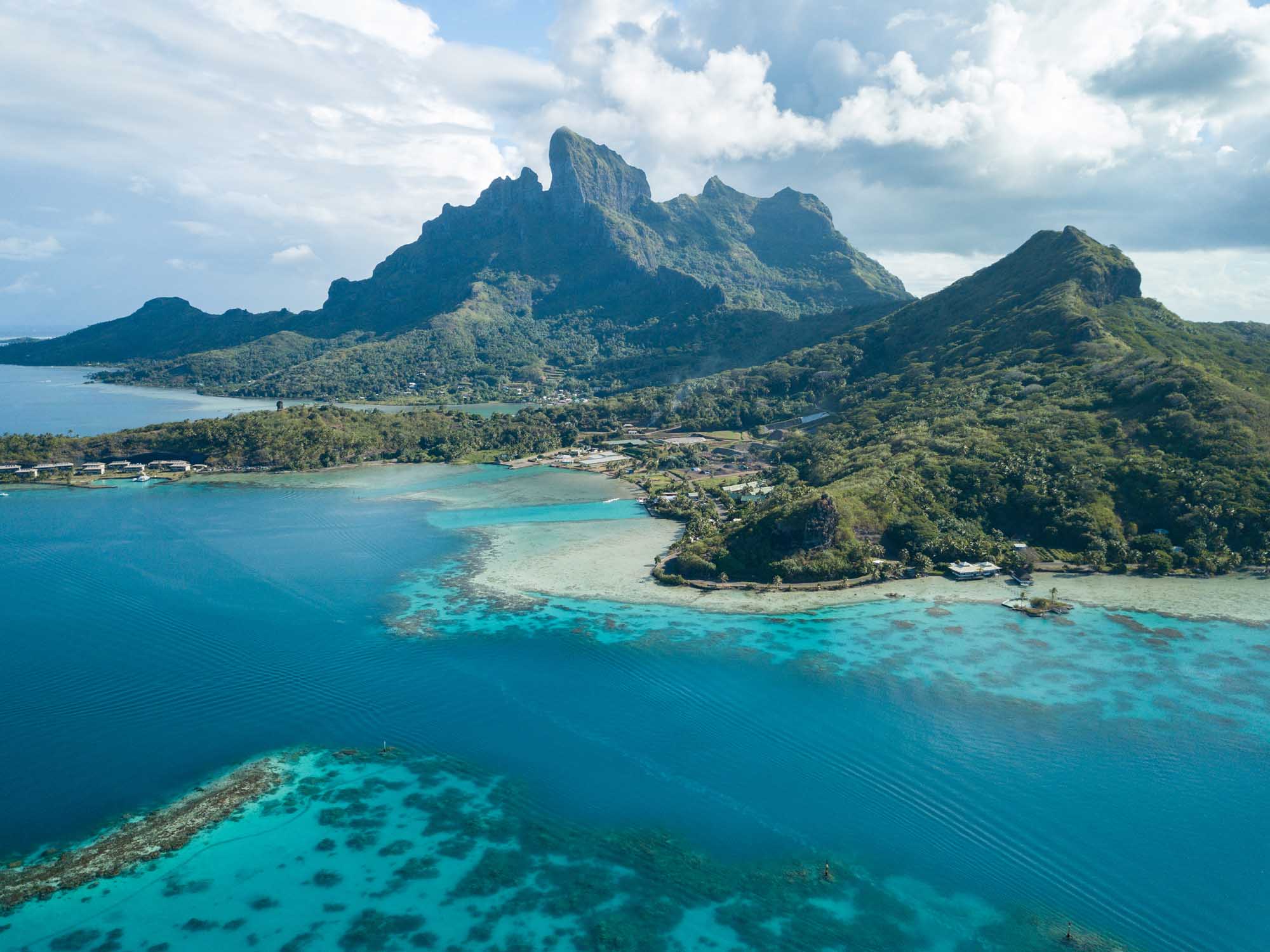An aerial drone photograph of Bora Bora island in French Polynesia, featuring the iconic Mount Otemanu rising dramatically from lush tropical vegetation, surrounded by stunning turquoise lagoon waters and coral reefs visible beneath the surface.