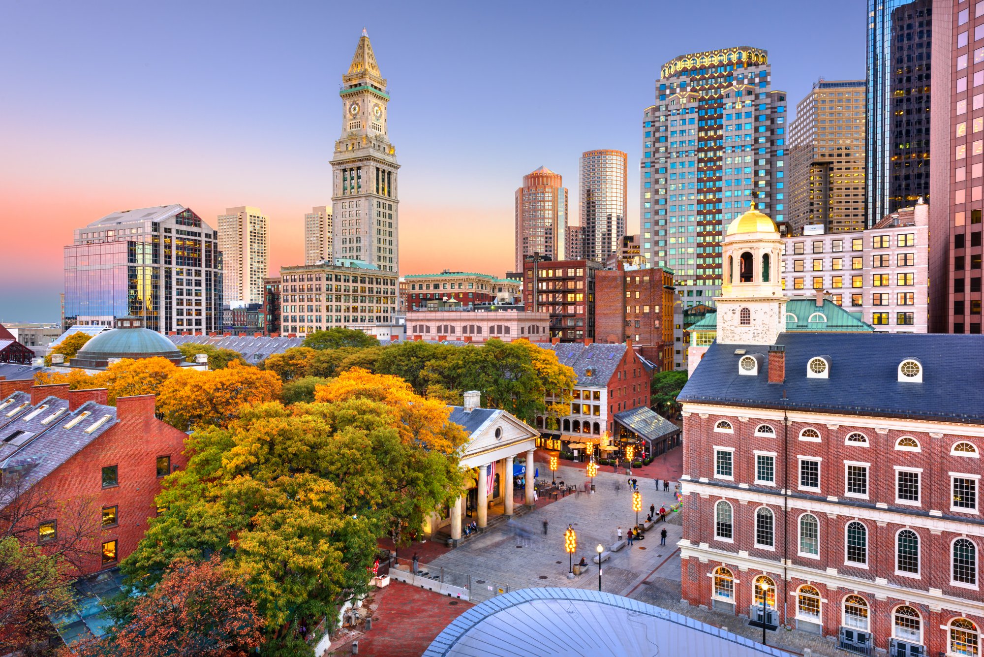 Boston Skyline with Historic Faneuil Hall A stunning autumn view of Boston's iconic skyline featuring the historic Faneuil Hall with its distinctive cupola in the foreground, surrounded by vibrant fall foliage, colonial-era brick buildings, and modern glass towers illuminated by the golden light of sunset.