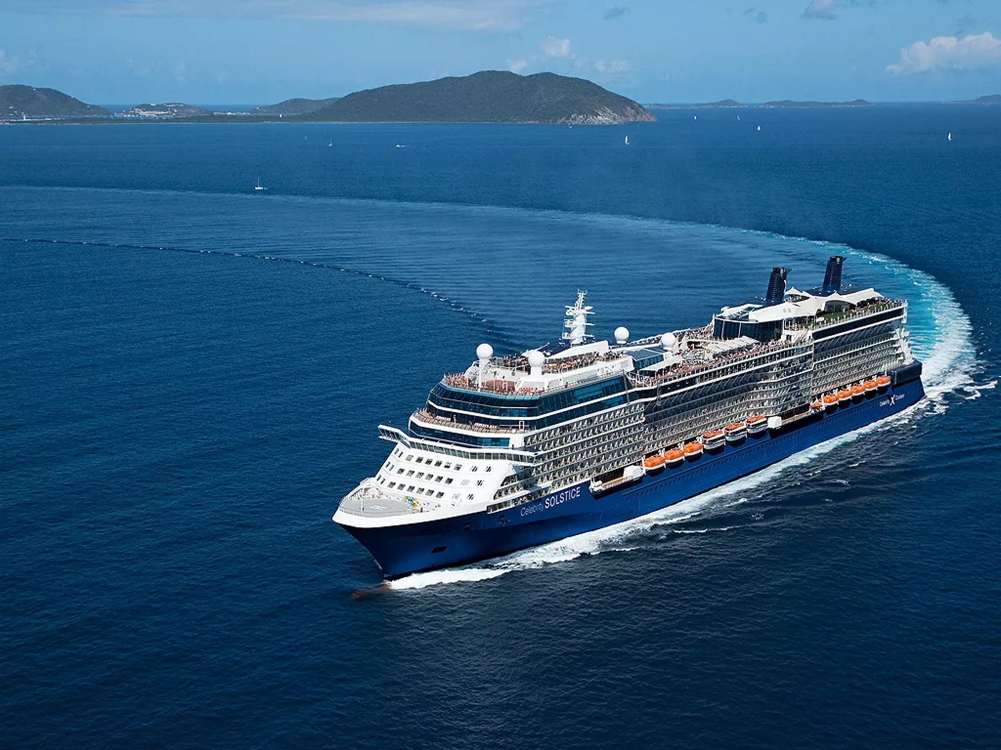 A large modern cruise ship with distinctive blue hull and orange lifeboats cruises through deep