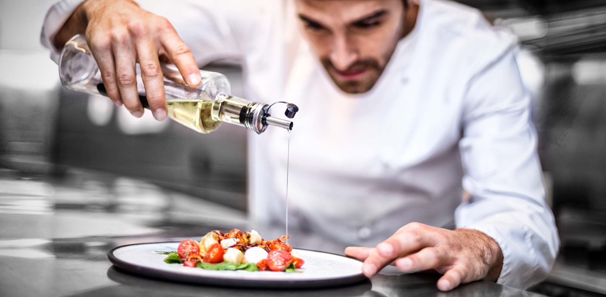 A professional chef in a white chef's coat carefully drizzles golden olive oil over an artfully plated dish featuring sliced tomatoes and other fresh ingredients in a commercial kitchen setting. The image captures a moment of culinary precision and fine dining preparation.