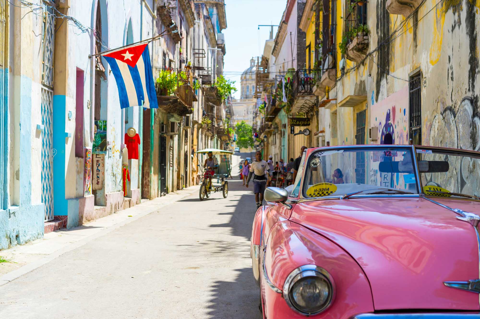 Classic Car and Cuban Flag in Havana Street A vibrant street scene in Havana, Cuba featuring a pink vintage American car parked along
