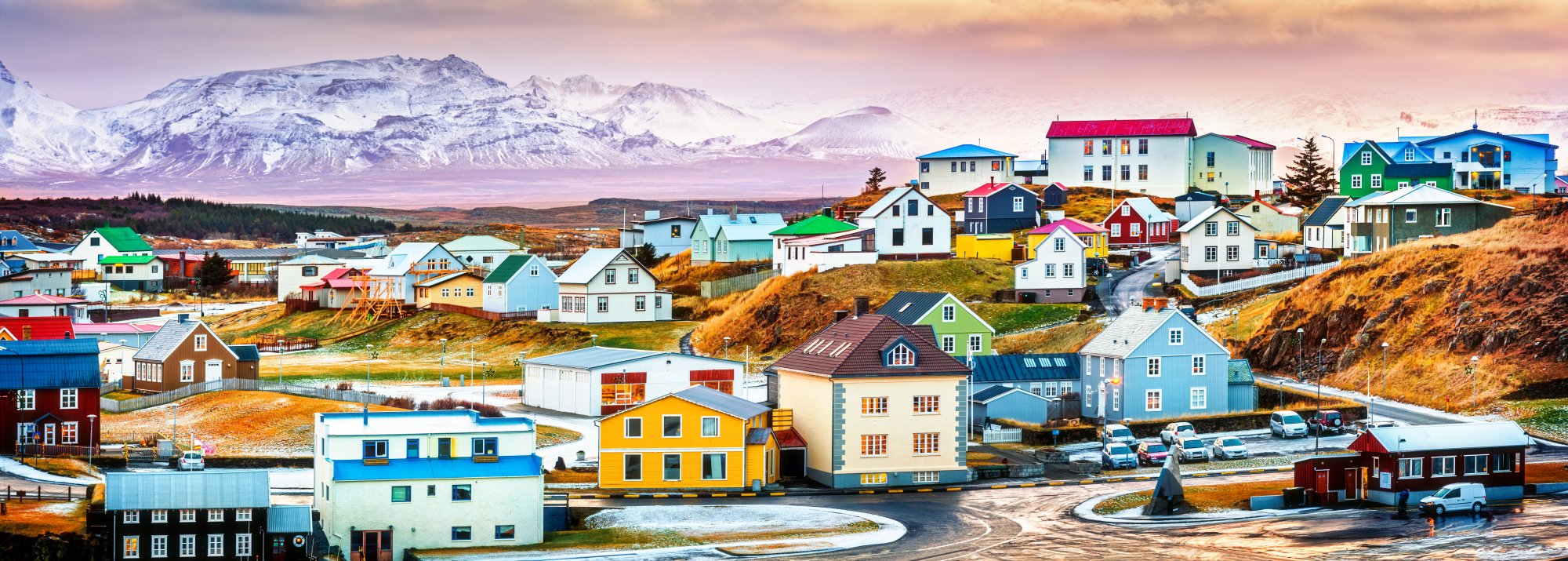 Colorful Arctic Village with Snowy Mountains A vibrant Greenlandic settlement featuring distinctive brightly-colored houses with grass-covered roofs arranged across rolling tundra, with a snow-capped mountain range dominating the background and small fishing boats visible in the harbor. The scene captures the unique Arctic architectural style and natural landscape characteristic of Greenland's remote communities.