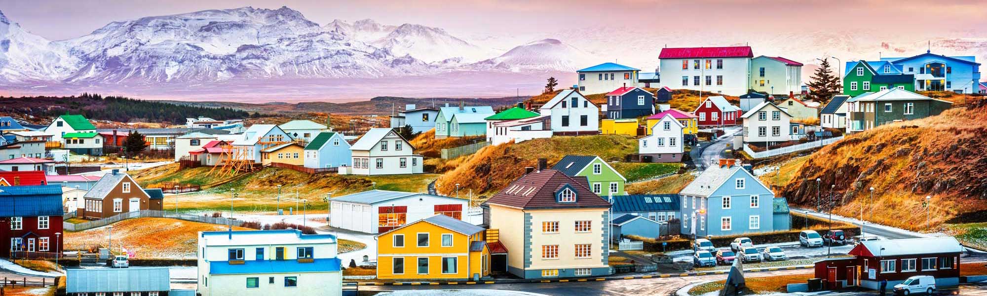Colorful Coastal Town in Arctic Landscape A vibrant Arctic coastal settlement featuring distinctively colorful houses in bright hues of blue, yellow, red, and green nestled against snow-capped mountains and moorland terrain. The picturesque village showcases traditional Nordic architecture with a mix of residential buildings, small boats, and vehicles arranged along a waterfront, characteristic of Greenlandic settlements.