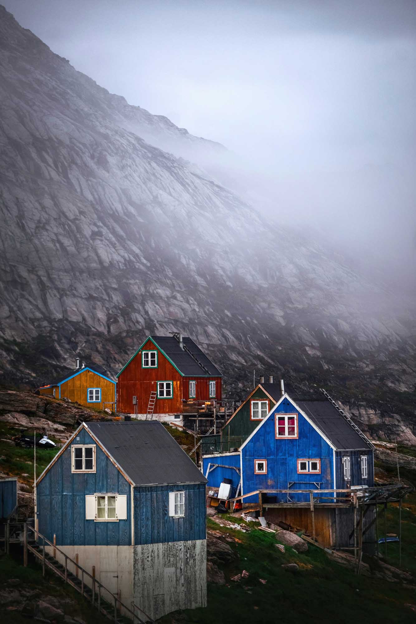 Colorful Houses Beneath Misty Arctic Cliffs A rustic orange-red cabin sits on a rocky Arctic shore overlooking a pristine fjord filled with icebergs and glaciers in the distance. The stark, barren landscape and massive ice formations exemplify the raw beauty of Arctic exploration and adventure travel.