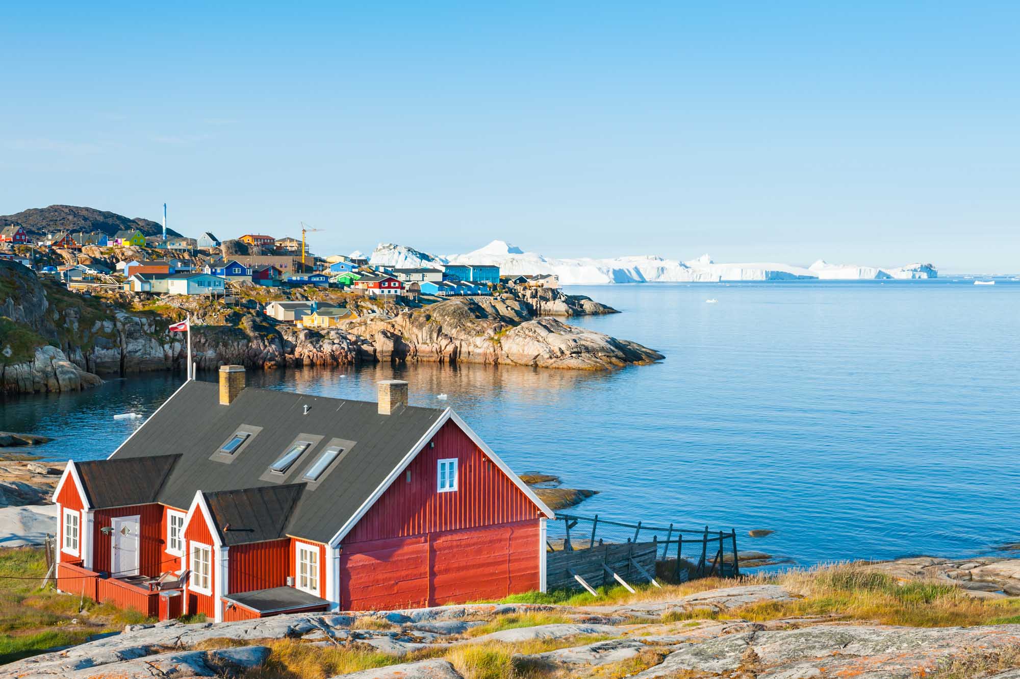 A picturesque red Greenlandic house sits on rocky terrain overlooking the Atlantic Ocean, with the vibrant settlement of Ilulissat visible across the bay and icebergs dotting the horizon. The scene captures the distinctive Arctic architecture and pristine coastal landscape characteristic of western Greenland.