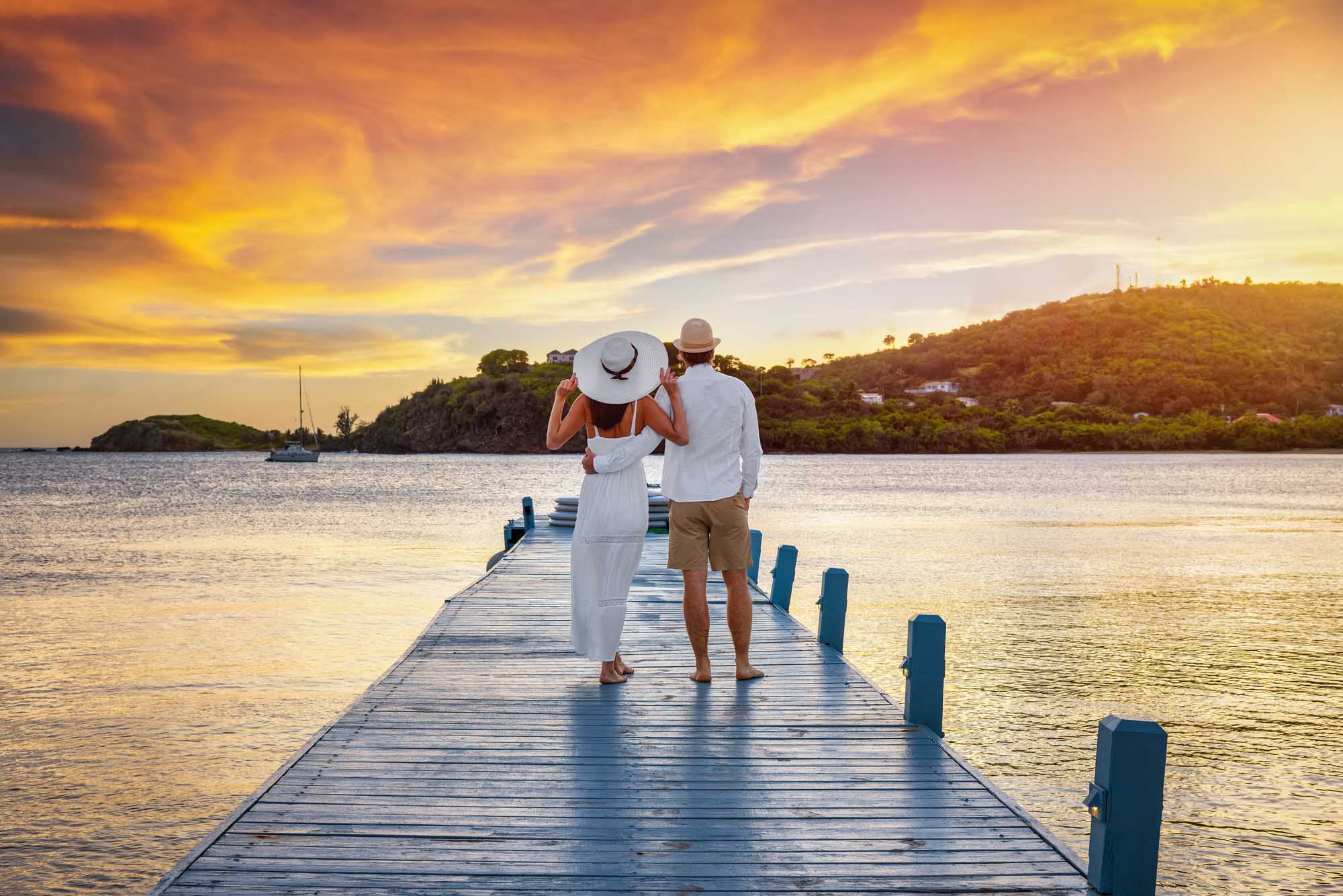 Couple on Pier at Tropical Sunset A romantic couple stands on a wooden pier overlooking calm tropical waters at golden sunset, with lush green islands and sailboats visible in the background. The woman wears a white flowing dress and wide-brimmed hat while the man wears light casual clothing, capturing an idyllic honeymoon or vacation moment in a Caribbean paradise setting.