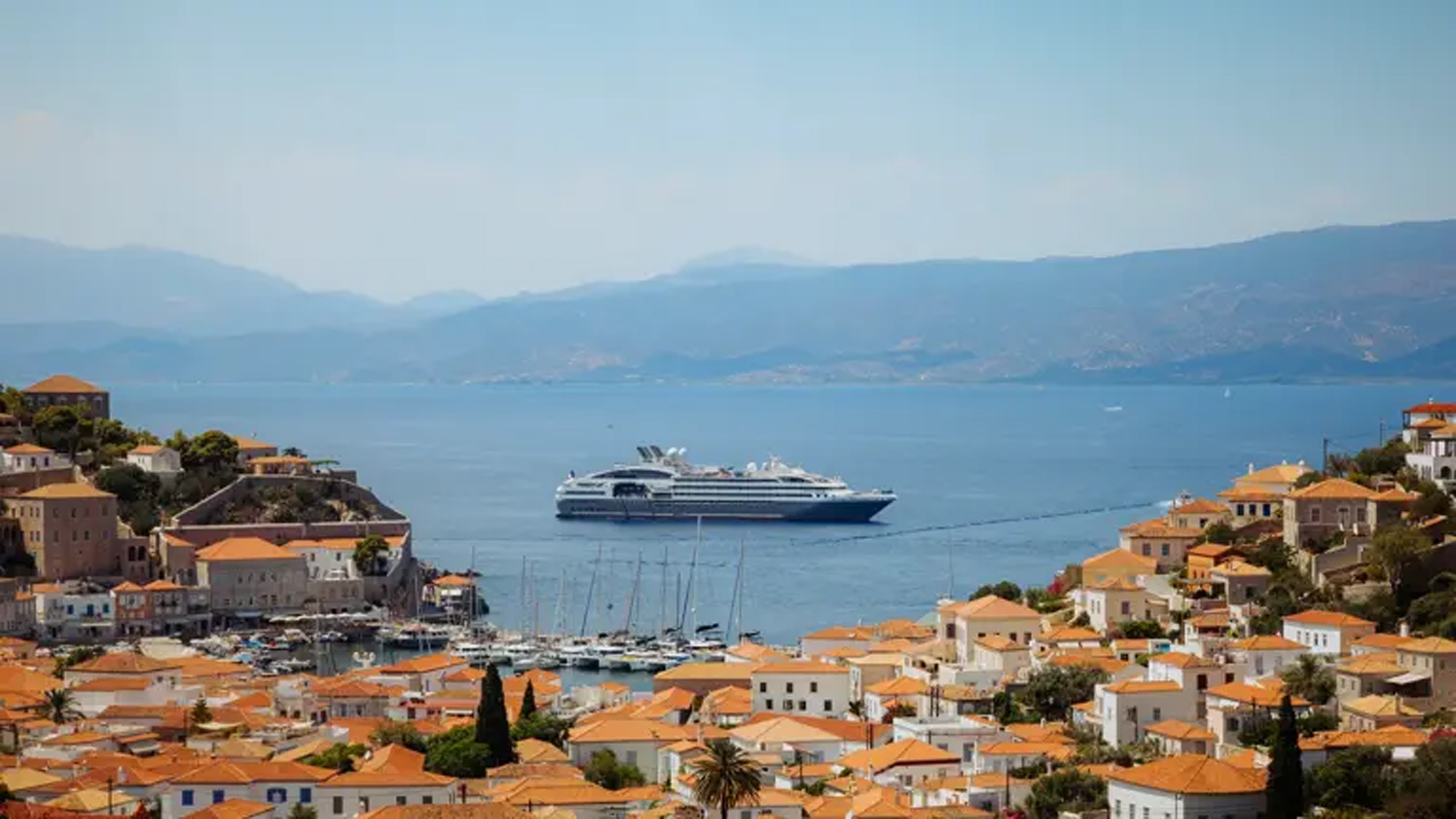 A large modern cruise ship anchored in the crystalline Mediterranean waters off the picturesque Greek island of Hydra, with traditional whitewashed buildings featuring orange-tiled roofs cascading down the hillside.