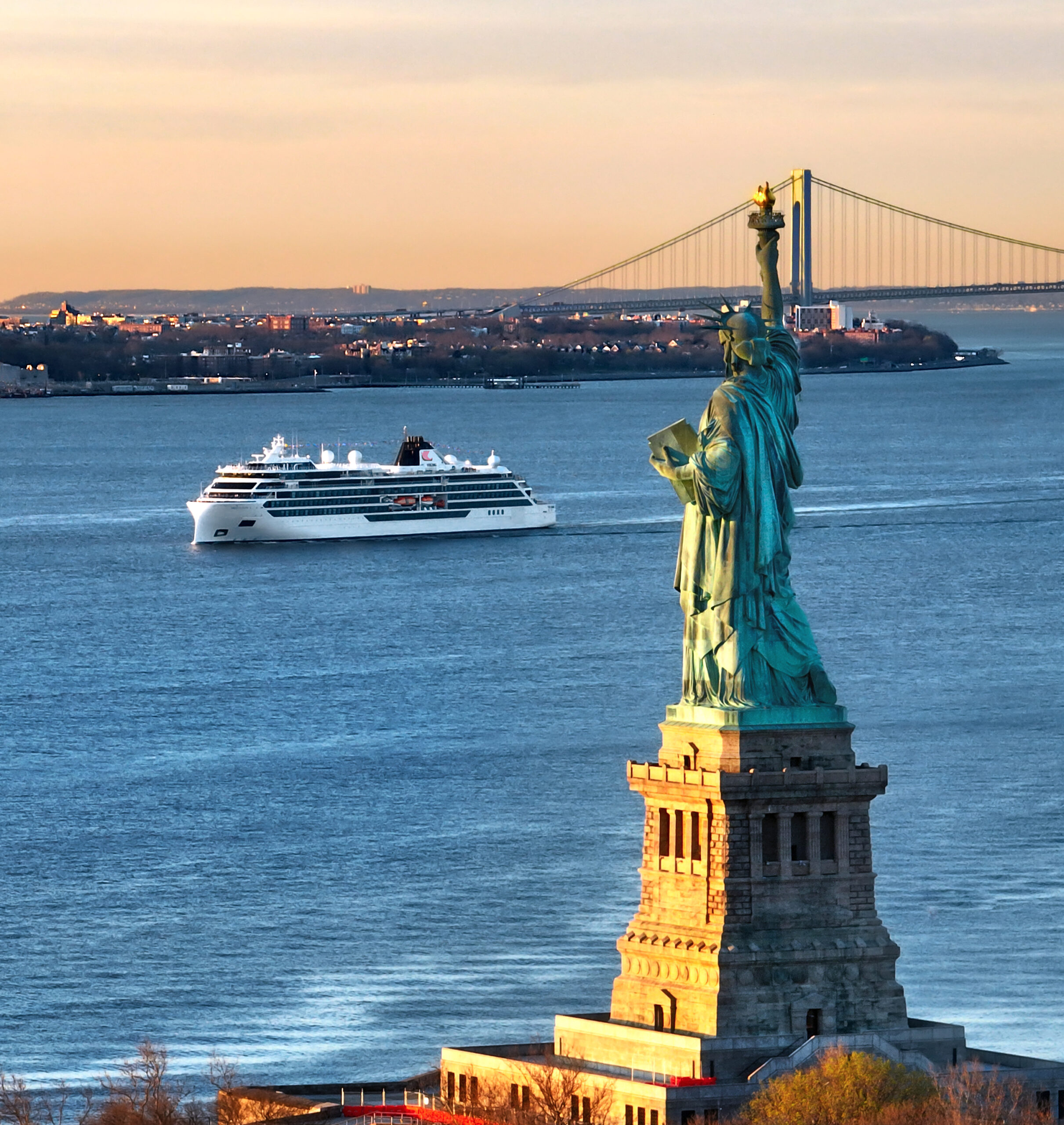 Cruise Ship Passing Statue of Liberty, New York A modern cruise ship enters New York Harbor past the iconic Statue of Liberty on Liberty Island, with the Verrazano-Narrows Bridge visible in the background and the Manhattan skyline dotted with buildings along the shoreline. The scene captures a golden-hour moment with warm sunlight illuminating the statue's copper patina and the ship's white hull against the blue harbor waters.