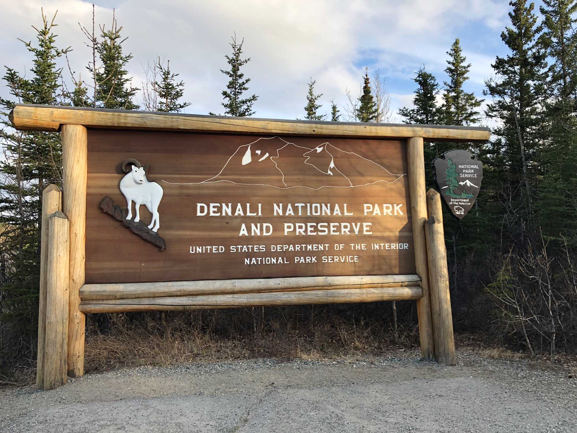 Denali National Park and Preserve Entrance Sign A rustic wooden entrance sign for Denali National Park and Preserve in Alaska, featuring a white Dall sheep illustration and mountain outline artwork. The sign is crafted from natural logs and displays official National Park Service branding, surrounded by boreal forest vegetation.