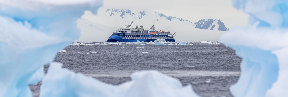A modern expedition cruise ship navigates through icy Antarctic waters, framed by massive icebergs and snow-covered peaks in the background. The vessel features distinctive blue hull livery and is positioned among pristine polar ice formations under a partly cloudy sky.