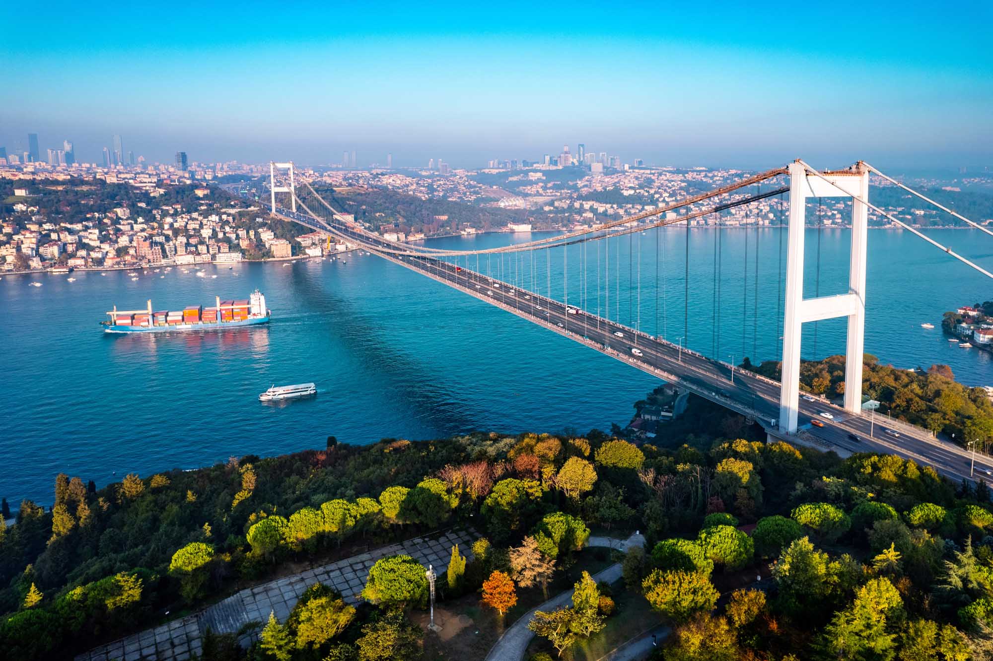 An aerial view of the iconic Fatih Sultan Mehmet Bridge spanning the Bosphorus Strait in Istanbul, Turkey, with a cargo ship and ferry navigating the turquoise waters below, the Asian coastline visible in the distance, and autumn-colored forests in the foreground.