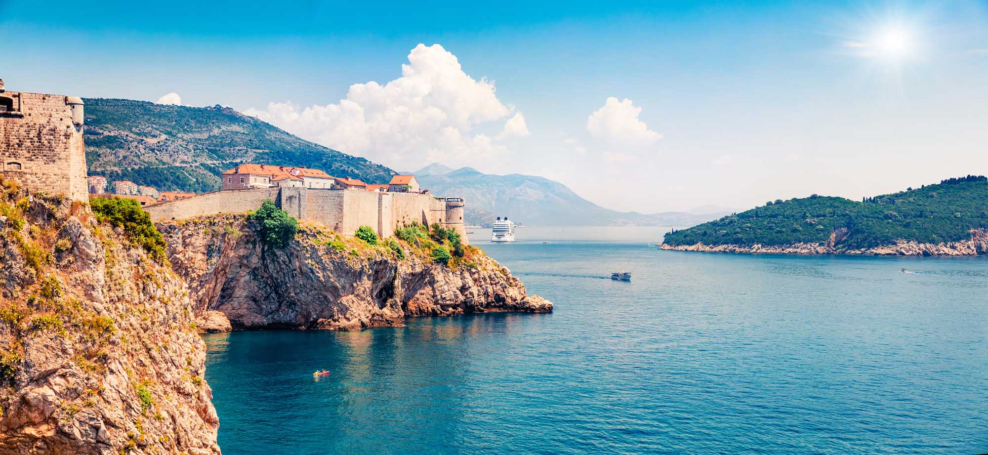 Fort Bokar Dubrovnik with Cruise Ship A panoramic view of the iconic Fort Bokar fortress perched on rocky cliffs in Dubrovnik, Croatia, overlooking the turquoise Adriatic Sea. A large cruise ship is visible in the harbor, with lush green islands and mountains in the background under a bright blue sky with white clouds.