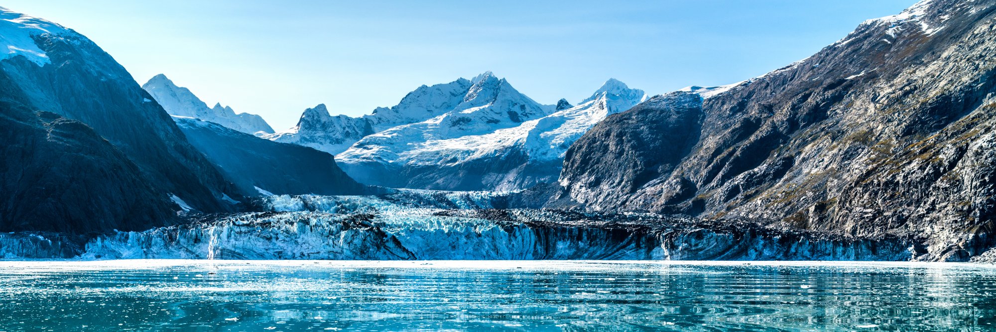 A stunning glacial landscape featuring a massive tidewater glacier flowing between snow-capped peaks into a turquoise-blue glacial lake, surrounded by steep mountain cliffs and rocky terrain under clear skies.