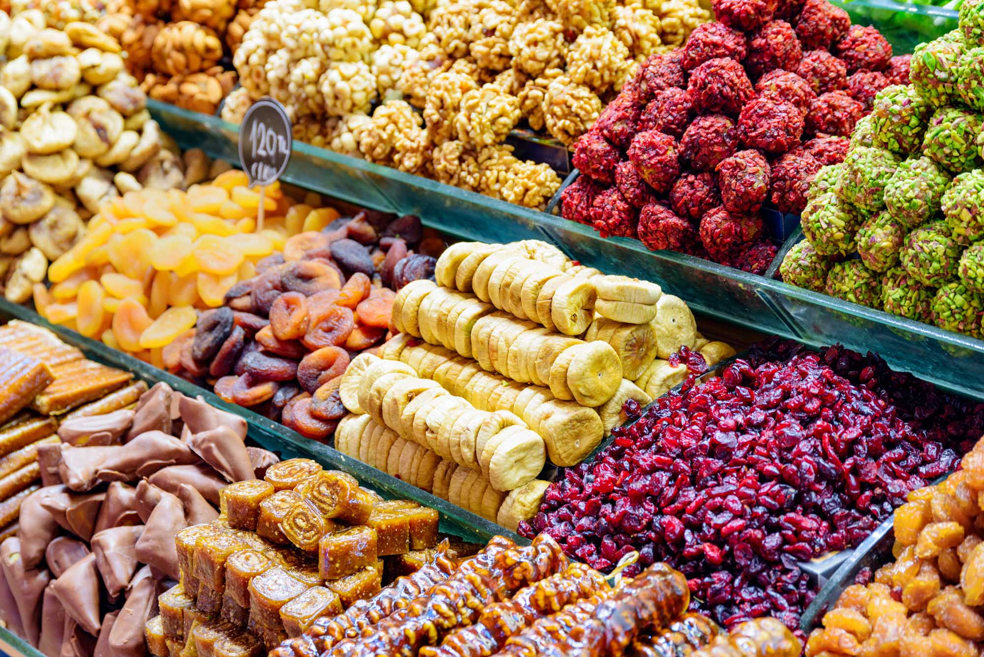 A vibrant display of traditional Turkish sweets and dried fruits at the Grand Bazaar in Istanbul. The market stall showcases an array of colorful confections including baklava, dried apricots, banana chips, fresh raspberries, mulberries, and various wrapped candies arranged in green display trays, representing the rich culinary heritage of this historic marketplace.